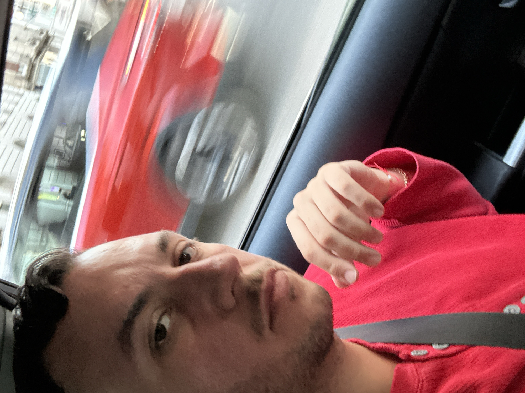 A selfie of a man in a red shirt wearing a seatbelt in a moving vehicle. Through the car window, a red double-decker bus is blurred, indicating the vehicle is in motion. The urban environment outside the window appears consistent with Madrid, Spain, due to the style of the bus and architecture.
