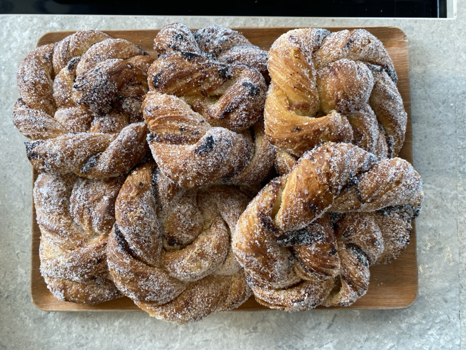 A rectangular wooden tray is positioned centrally, containing multiple golden-brown, braided pastries. Each pastry is densely coated with white granulated sugar. Sporadic dark flecks, consistent with chocolate or cinnamon filling, are visible within the twisted dough. The tray rests on a light grey, subtly textured surface, indicative of a countertop. A dark, undefined object is partially visible along the right edge of the frame. No individuals, actions, or specific interactions are observed. The scene depicts an indoor environment, likely a kitchen or bakery. The precise location within Tel-Aviv, Israel, cannot be determined from the visual information provided.