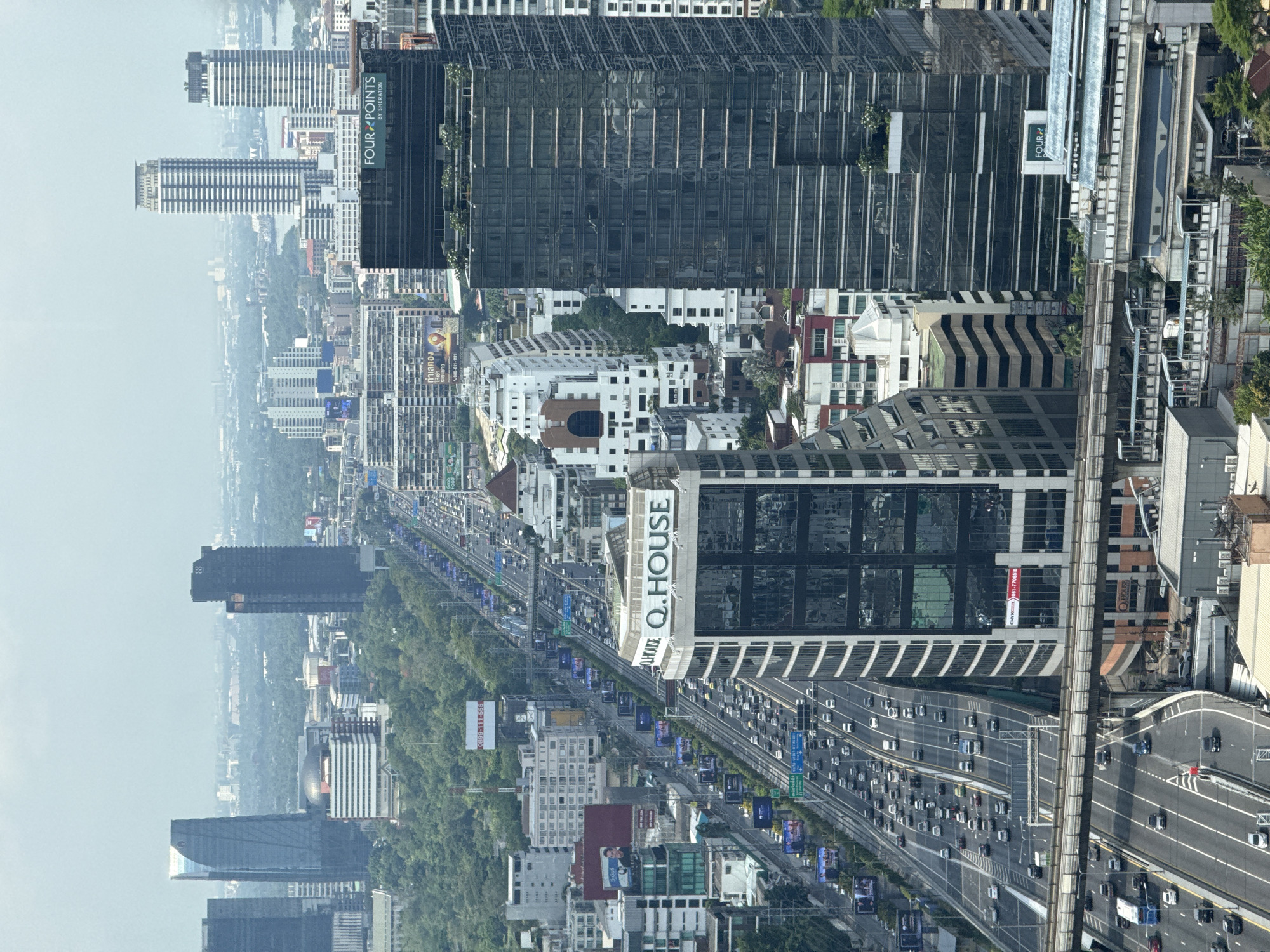 This is an aerial view of Bangkok, Thailand. The image captures a densely populated urban landscape with a prominent highway running through the center. The highway is congested with vehicular traffic, suggesting a busy time of day. The skyline features numerous high-rise buildings, including the "Four Points by Sheraton" and "Q.House" skyscrapers. The architectural styles of the buildings vary, showcasing a mix of modern and older structures. Lush greenery interspersed among the buildings indicates the presence of parks or gardens within the city. The sky appears overcast, casting a soft light over the scene.
