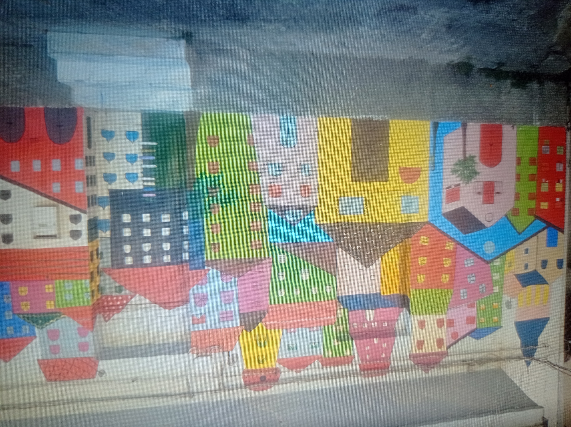 A vibrant, colorful mural depicting a whimsical cityscape adorns an outdoor wall. The artwork features a collection of densely packed, cartoon-like buildings with steeply pitched roofs and numerous windows. The buildings are painted in a variety of bright hues, including yellow, pink, green, blue, and red, with accents of white and brown. Some buildings have distinctive architectural details, such as cupolas, arched windows, and chimneys. In the foreground, a rough-textured wall separates the mural from a paved area, possibly a street or walkway, where a set of concrete steps leads upwards. The overall impression is one of childlike fantasy and playful design. There are no people or animals visible in the mural or the surrounding environment. The mural is located in the city of Mukachevo, Ukraine.