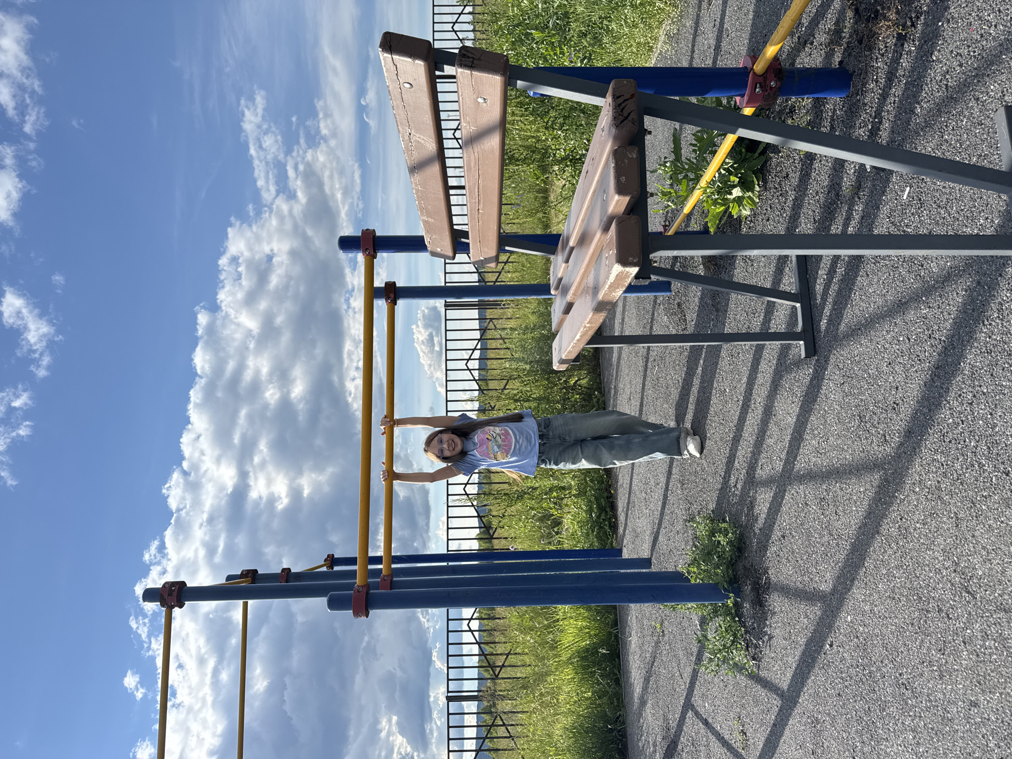 In Mursalimkino, Russia, a young girl is captured in a playful moment on a playground. She is hanging from a horizontal bar, her arms extended and a cheerful expression on her face, suggesting a sense of joy and freedom. She wears a light blue t-shirt with a graphic design and baggy green pants, paired with white sneakers.
The playground equipment features blue and yellow bars, some connected by red clamps, suggesting it's a sturdy structure. A wooden bench with weathered planks is visible in the background, hinting at the playground's usage. The ground is a dark, textured asphalt, with patches of green grass and weeds growing around the bases of the equipment, indicating an outdoor setting that experiences natural growth.
The scene is set against a backdrop of a bright blue sky filled with dramatic, puffy white clouds, characteristic of a pleasant day. The lighting suggests it's daytime, likely midday or afternoon, with distinct shadows cast by the playground equipment on the asphalt. The overall atmosphere is relaxed and summery, evoking a sense of childhood fun and outdoor activity. There is no visible text in the image.