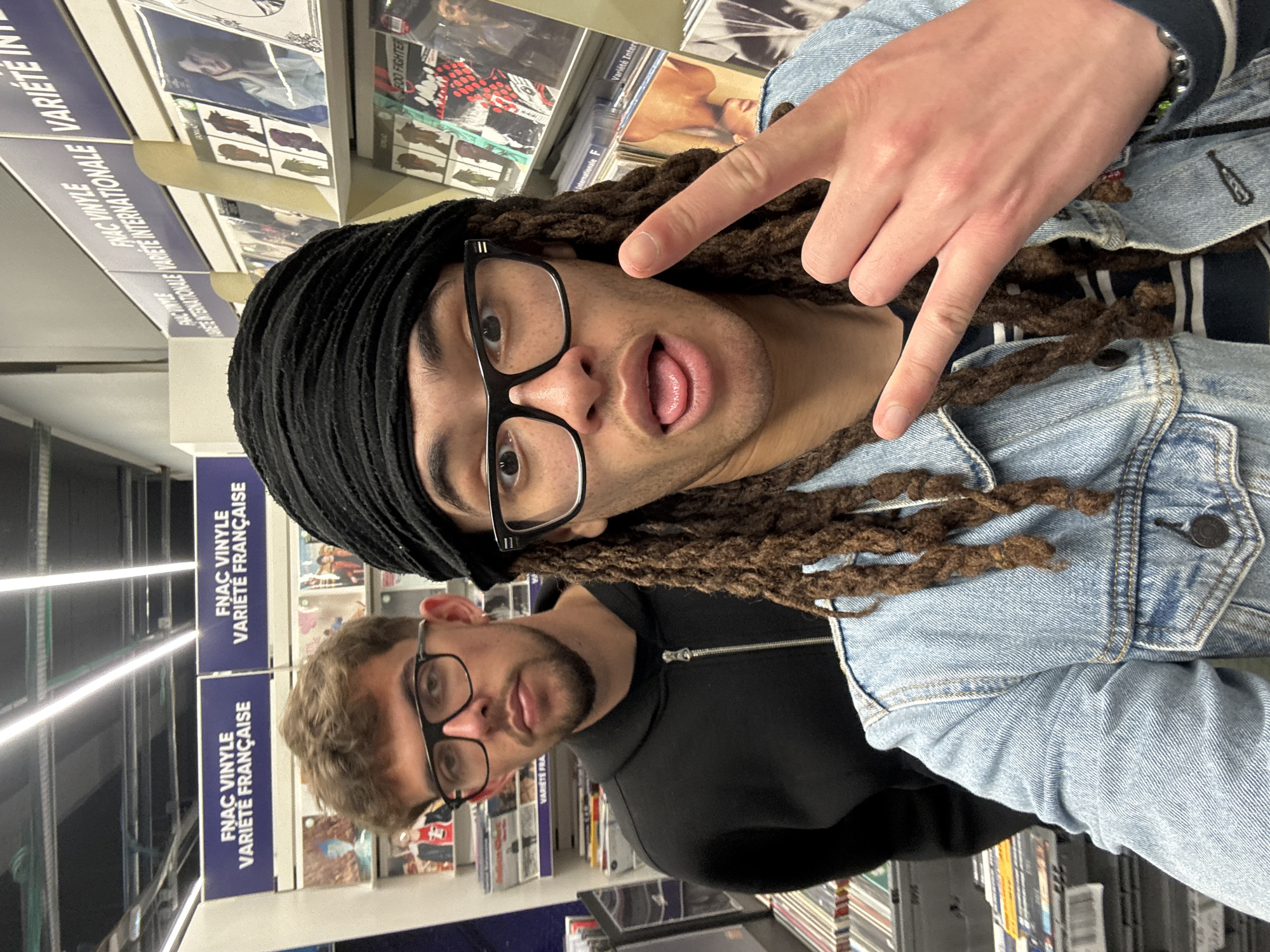 The image shows two individuals in what appears to be the FNAC store in Villebon-sur-Yvette, France. The setting includes racks of vinyl records and related signage. The first person, positioned in the foreground, wears a black head covering, glasses, and a denim jacket. They are sticking their tongue out and making a "peace sign" with one hand. The second person, partially visible behind the first, wears glasses and a black hooded sweatshirt.