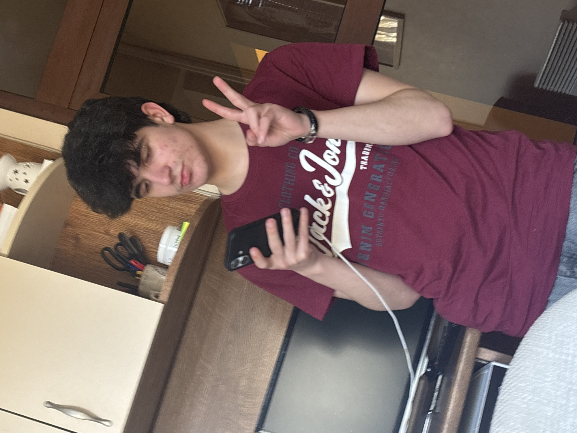 A young male with dark hair is in an interior setting, possibly a kitchen. He is wearing a maroon t-shirt with a "Jack & Jones" logo. He is holding a black smartphone in his left hand and making a peace sign with his right hand. A white charging cable is connected to the phone. Behind him are cabinets and countertops, which contain objects, including scissors and cups. The image's location in Pleven, Bulgaria, cannot be confirmed from the visual data.
