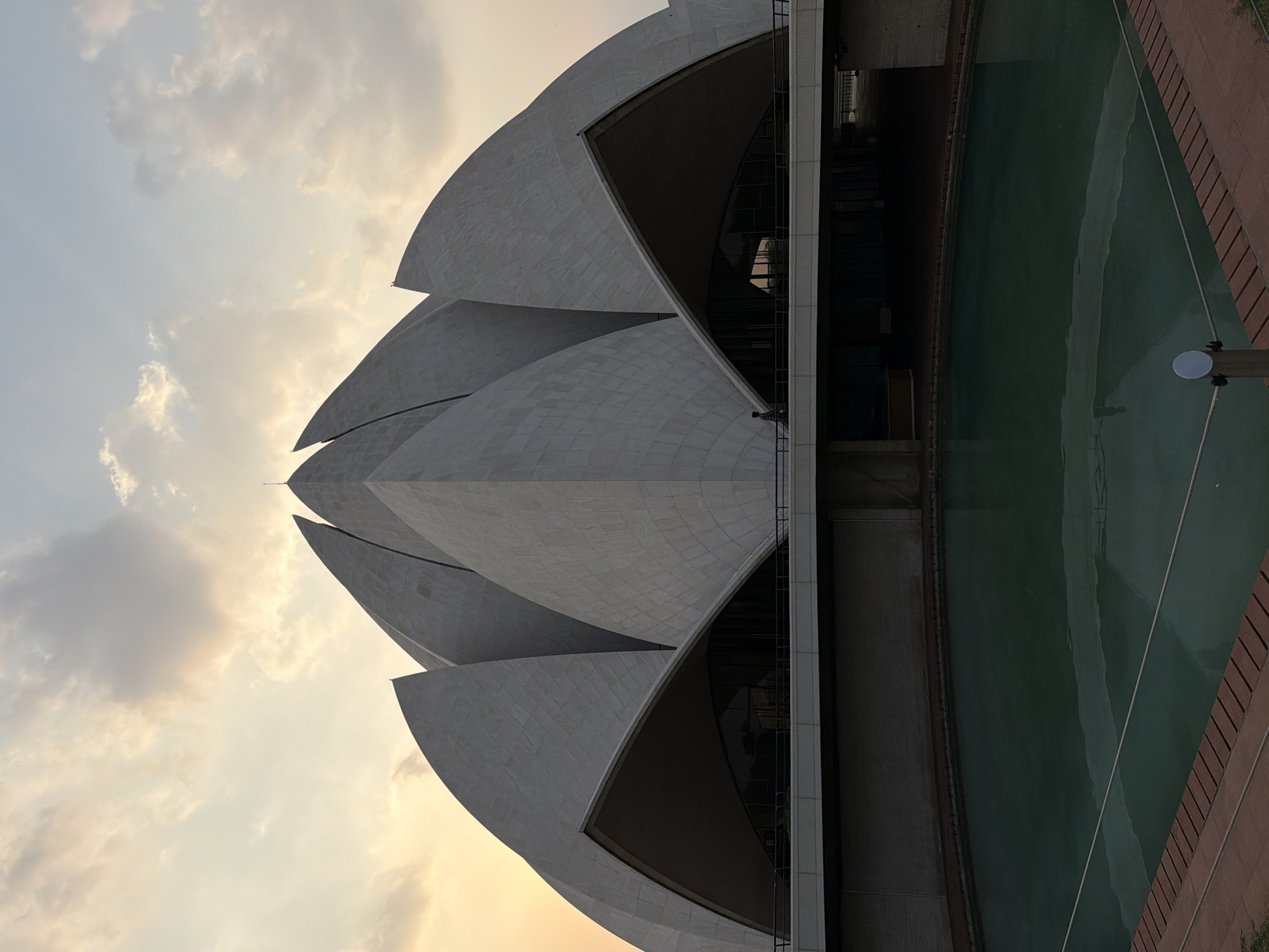 The image, rotated 90 degrees clockwise, prominently features the unique architecture of the Lotus Temple in Delhi, India, under a softly lit, partially cloudy sky. The iconic structure, designed to resemble a half-opened lotus flower, dominates the right side of the frame. Its distinct white marble or concrete 'petals' are visible, gracefully curving upwards and outwards. Some of the petals are illuminated by the warm light, while others are in shadow, creating depth and contrast. Dark, recessed areas beneath the petals likely indicate glass windows or entrances.
To the bottom right of the temple, a rectangular reflecting pool with dark greenish-blue water is visible, bordered by a reddish-brown walkway or ground. The water surface shows subtle ripples and reflections. The sky, occupying the upper left portion of the image, displays a blend of light blue and soft orange/yellow hues, suggesting either dawn or dusk. Scattered, wispy clouds are present, adding texture to the sky. The overall atmosphere is serene and majestic, highlighting the architectural beauty of the landmark during a golden hour. There is no visible text in the image.