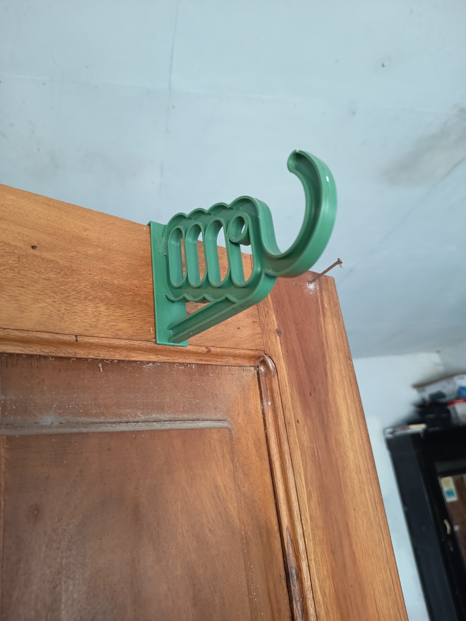 A close-up shot shows a green plastic hook attached to the top edge of a wooden door frame. The hook has a decorative, scalloped edge along its base and extends outward in a curved shape. A small nail is visible protruding from the wooden frame near the hook. The background consists of a light blue wall. The image appears to be taken indoors, possibly in a residential setting. There are no people or actions depicted in the image.