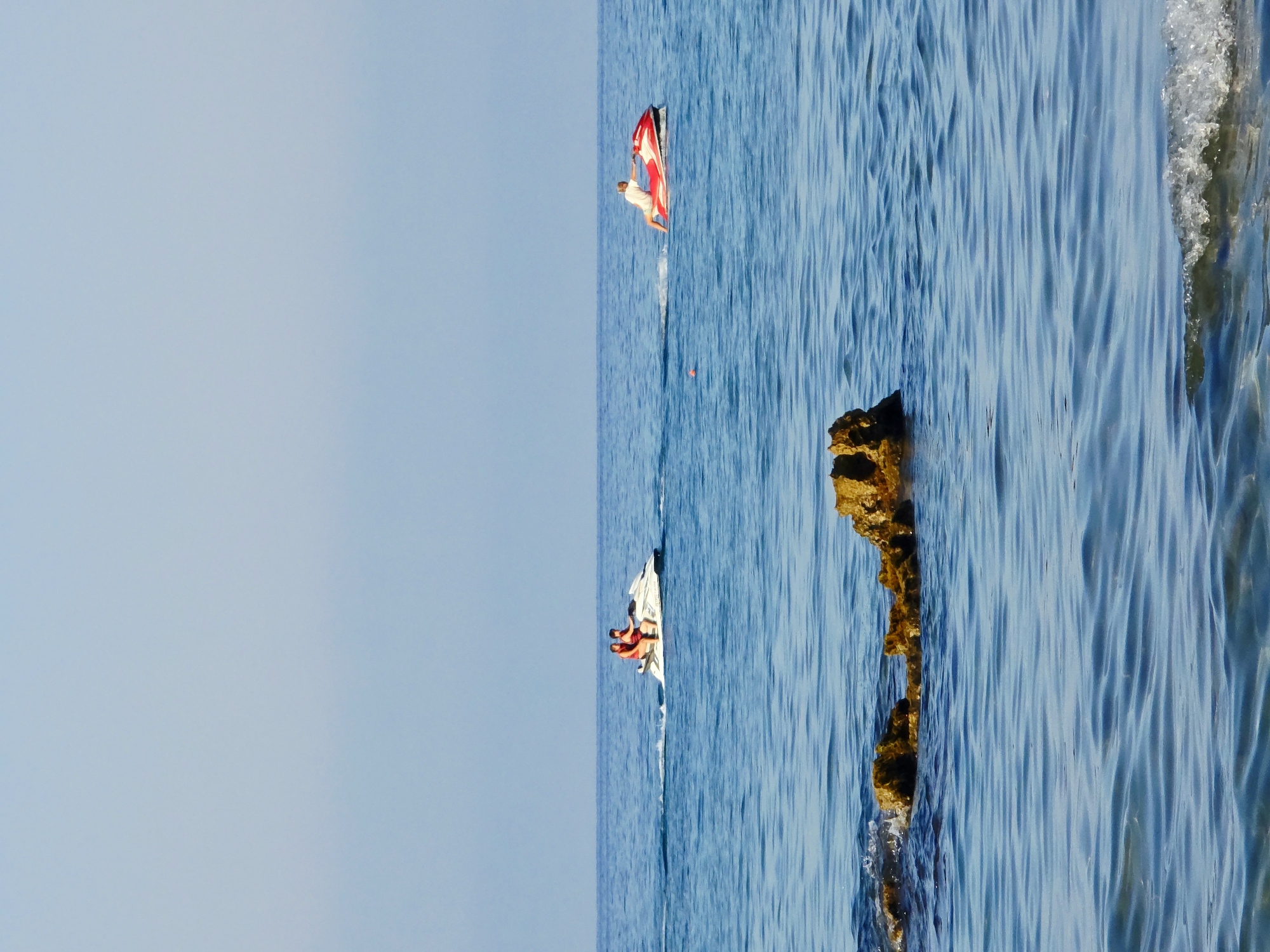 A horizontal, eye-level shot depicts a serene seascape with two jet skis navigating the calm, blue waters under a clear, pale blue sky. In the foreground, a cluster of rocks emerges from the water, breaking the surface. The mid-ground features a white jet ski with two individuals wearing orange life vests, moving from left to right. Further to the right, a red and white jet ski is ridden by a solitary individual in a white top. The jet skis are creating white wakes as they move across the water. The location appears to be a coastal area, consistent with the vicinity of Malia Municipal Unit, Greece, known for its beaches and water activities. The overall scene conveys a sense of leisure and outdoor recreation.