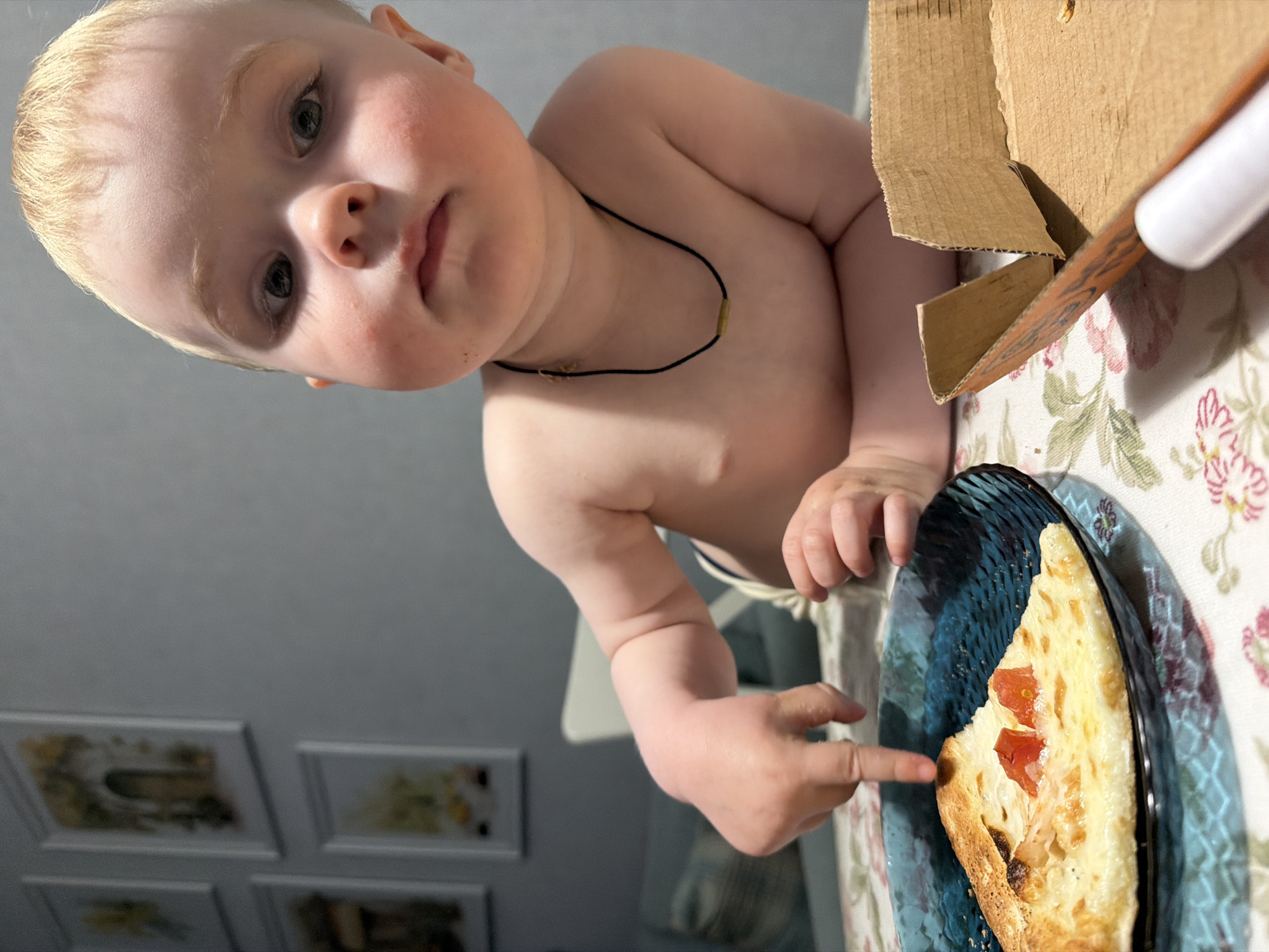 A shirtless fair-skinned child with blond hair and a black necklace is seated at a table with a floral patterned tablecloth. The child is reaching with their right hand towards a slice of pizza on a blue textured plate. A partially opened cardboard box and a knife are on the table near the plate. In the background, framed pictures are visible on a light gray wall. There is no evidence to suggest the location of the photo is in Votkinsk, Russia.