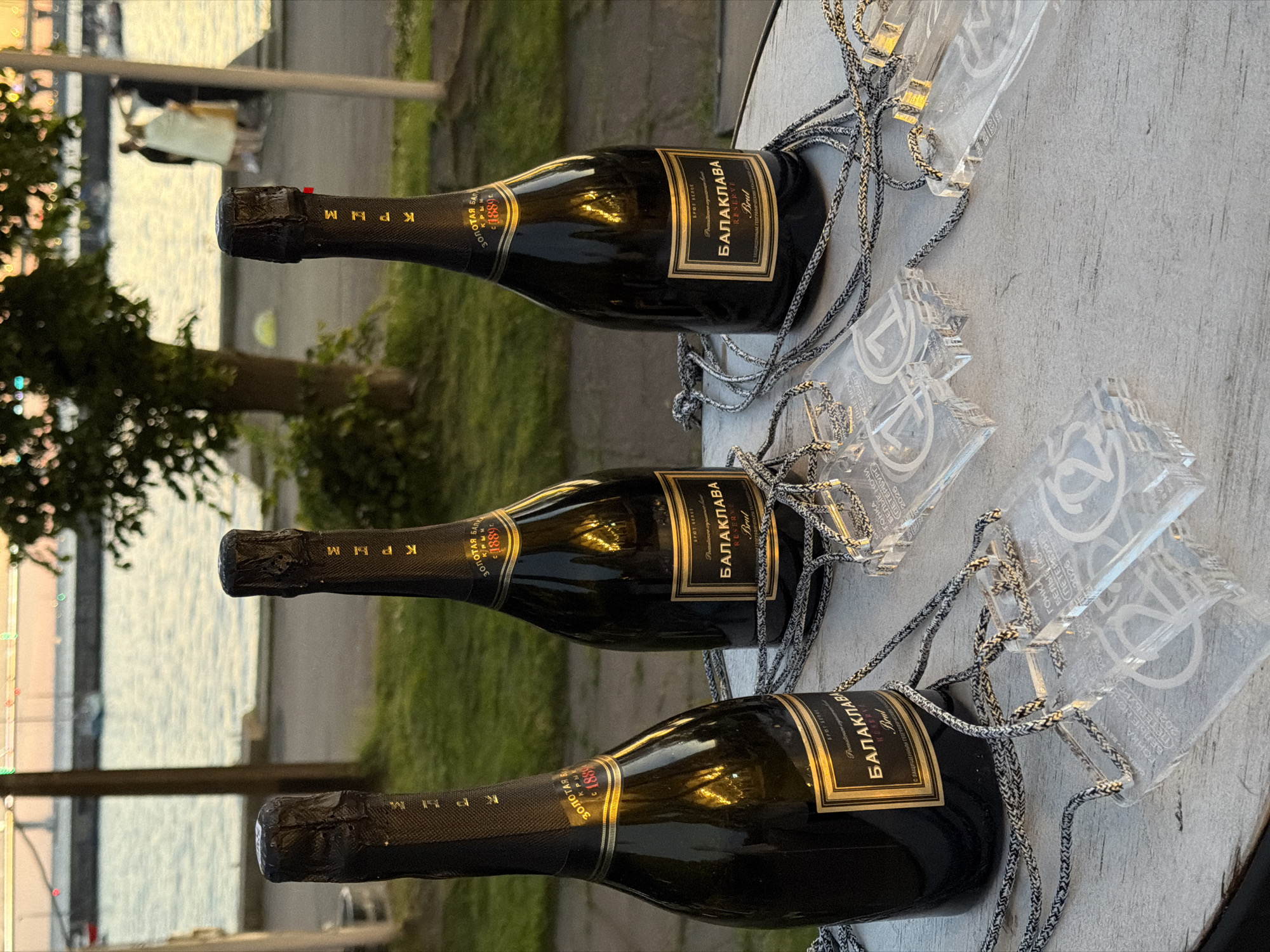 Three bottles of black champagne with gold labels are arranged on a rustic wooden table. Each bottle has a foil cap. Attached to the bottles are clear acrylic pendants with silver lanyards, which appear to be awards or commemorative items. The background suggests an outdoor setting, possibly a waterfront with a body of water and a distant shoreline. In the far background, several people are visible, suggesting a gathering or event. The lighting indicates it is either dawn or dusk. The text on the champagne labels reads "ЗОЛОТАЯ БАЛКА" (Zolotaya Balka) and "БАЛАКЛАВА" (Balaklava), indicating a Crimean wine brand. The acrylic pendants have text that includes "ГОНКА БЕЛЫЕ НОЧИ" (Race White Nights) and "2025", strongly suggesting the location is Saint Petersburg, Russia, due to the "White Nights" cultural reference and the year displayed.