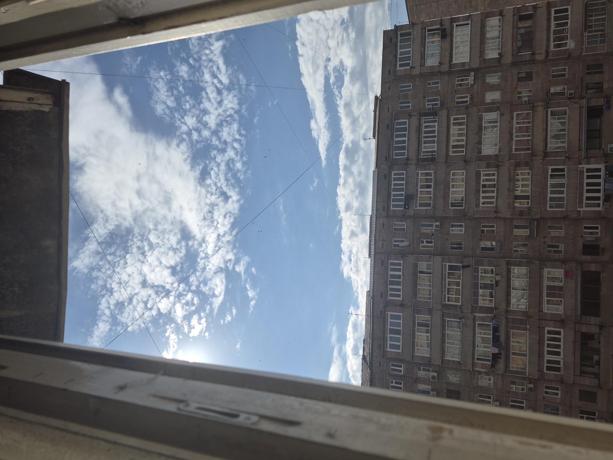 The image captures an upward view from an interior space, likely a window or balcony, framing a vibrant blue sky and a tall residential building. In the foreground, parts of a window frame or balcony railing, made of light-colored wood or metal, occupy the top and bottom edges, slightly obscuring the scene.
The sky, dominating the left and central portions of the image, is a clear blue adorned with scattered white, fluffy clouds, resembling cumulus or stratocumulus formations. The sun is visible as a bright flare in the bottom-left corner of the sky, indicating bright daylight conditions, possibly mid-morning or late afternoon. Several thin, dark utility wires crisscross the sky, and tiny dark specks, presumed to be birds, are visible against the blue.
On the right side of the frame, a large, multi-story apartment building stretches upwards. Its facade is a dull, uniform color, likely concrete or similar material, with numerous rectangular windows of varying sizes, many featuring white frames. Some windows or balconies show signs of daily life, such as hanging laundry. The architectural style appears utilitarian, characteristic of large residential blocks found in urban areas.
No explicit human activity is depicted, but the building suggests a busy residential environment. The scene is set in Yerevan, Armenia, offering a glimpse of an everyday urban landscape under a sunny sky. There is no visible text in the image.