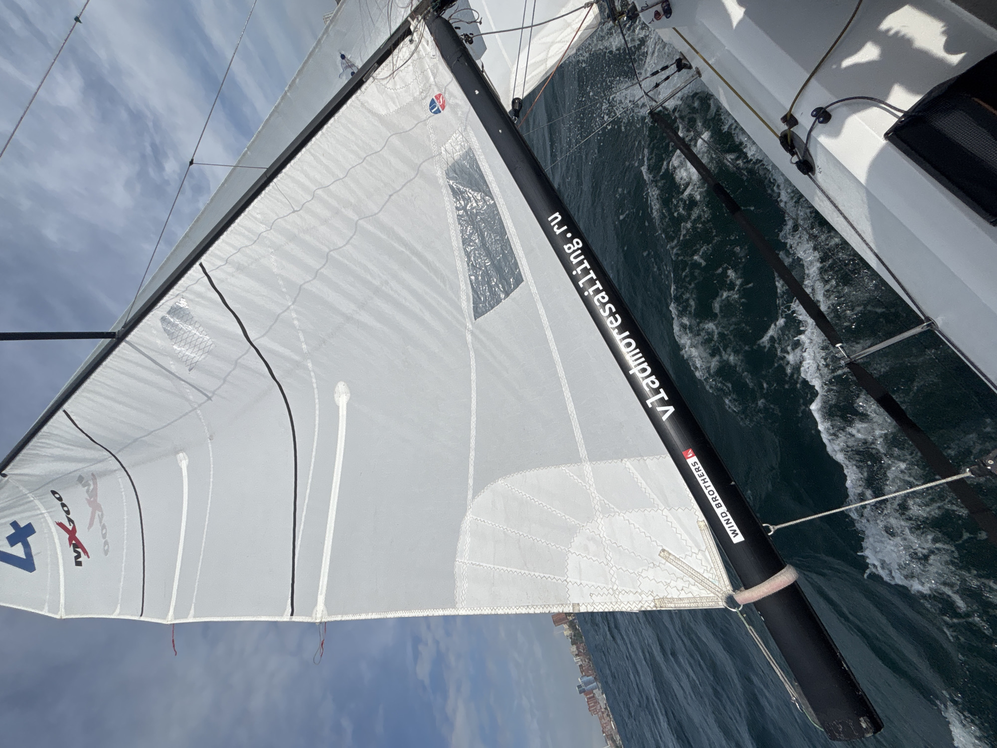 The image captures a sailboat on the water in Vladivostok, Russia. The angle suggests the photograph was taken from on board the vessel, with a view up at the sail and over the water. The sail is white, with visible brand markings and construction details. A black mast is prominent in the foreground, adorned with the text "Vladmorsailing.ru" and "WIND BROTHERS." The water shows signs of the boat's movement, indicated by the frothy wake. Along the horizon, buildings suggest the coastline of Vladivostok. The sky is partly cloudy.