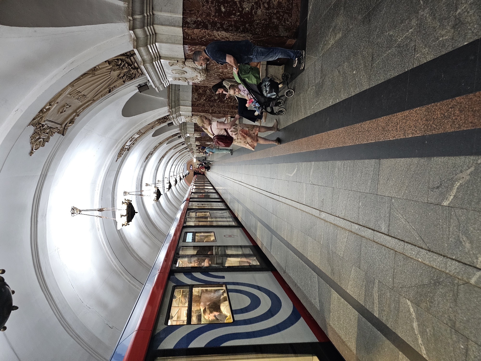The image depicts a subway platform in Moscow, Russia. The scene is captured with a perspective that highlights the tunnel's curvature and the train. People are present on the platform, including a man and a woman with a stroller, and another woman walking. The train itself is visible, with passengers visible through the windows. The architectural details of the subway station are prominent, including the arched ceiling, decorative elements, and lighting fixtures. The platform surface is composed of different shades of grey stone with a longitudinal dark grey and orange stripe.