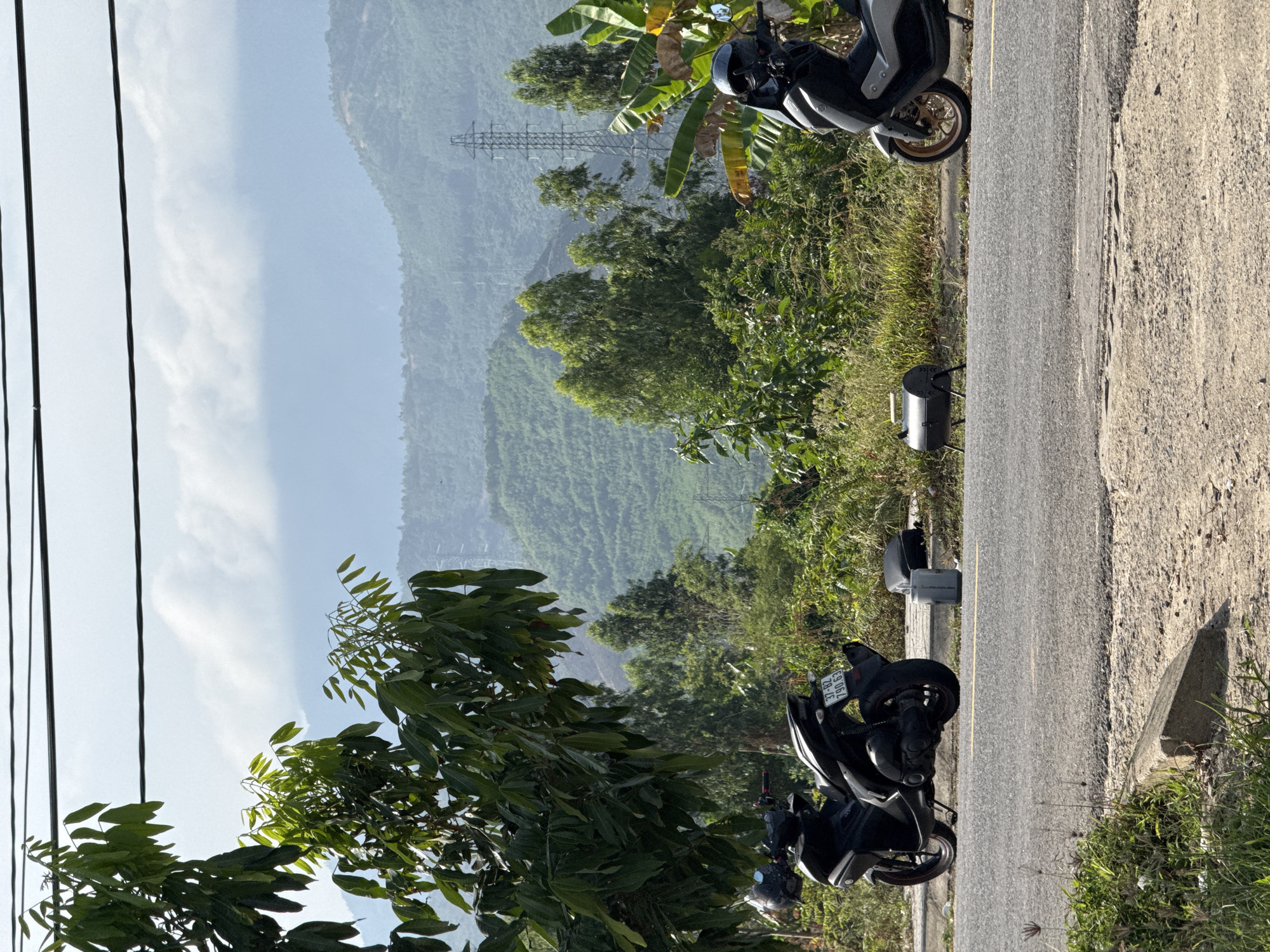 The image shows a scene along a road in a mountainous area, possibly within or near Đà Nẵng, Vietnam. The road is paved and lined with vegetation on the side. Several parked motorbikes are visible along the edge of the road, some partially obscured by foliage. A utility pole with power lines is visible to the left. In the background, a mountain range is visible, covered with dense green vegetation. Power transmission towers are visible on the hillside. The sky is hazy, suggesting a humid or overcast day.