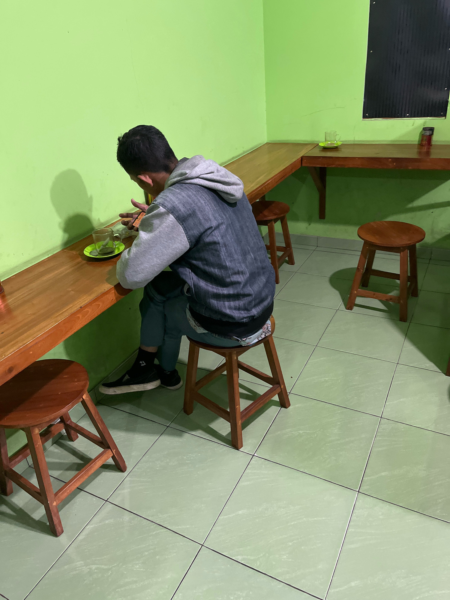 A young man with dark hair is seated at a wooden counter in a restaurant with lime green walls. He is wearing a gray hooded jacket over a dark shirt, blue jeans, and black sneakers with white soles. He is holding what appears to be a mobile phone or a small electronic device in his right hand. In front of him on the counter is a clear glass mug with a green coaster underneath it. The counter is L-shaped, extending to the right, and has two other wooden stools placed beneath it. In the background, a dark, rectangular object is mounted on the wall. The floor is tiled in a light green color with black grout lines. The lighting is dim, casting shadows from the stools and the man onto the walls and floor. The overall scene suggests a casual dining establishment in Banguntapan, Indonesia, possibly a local eatery or cafe.