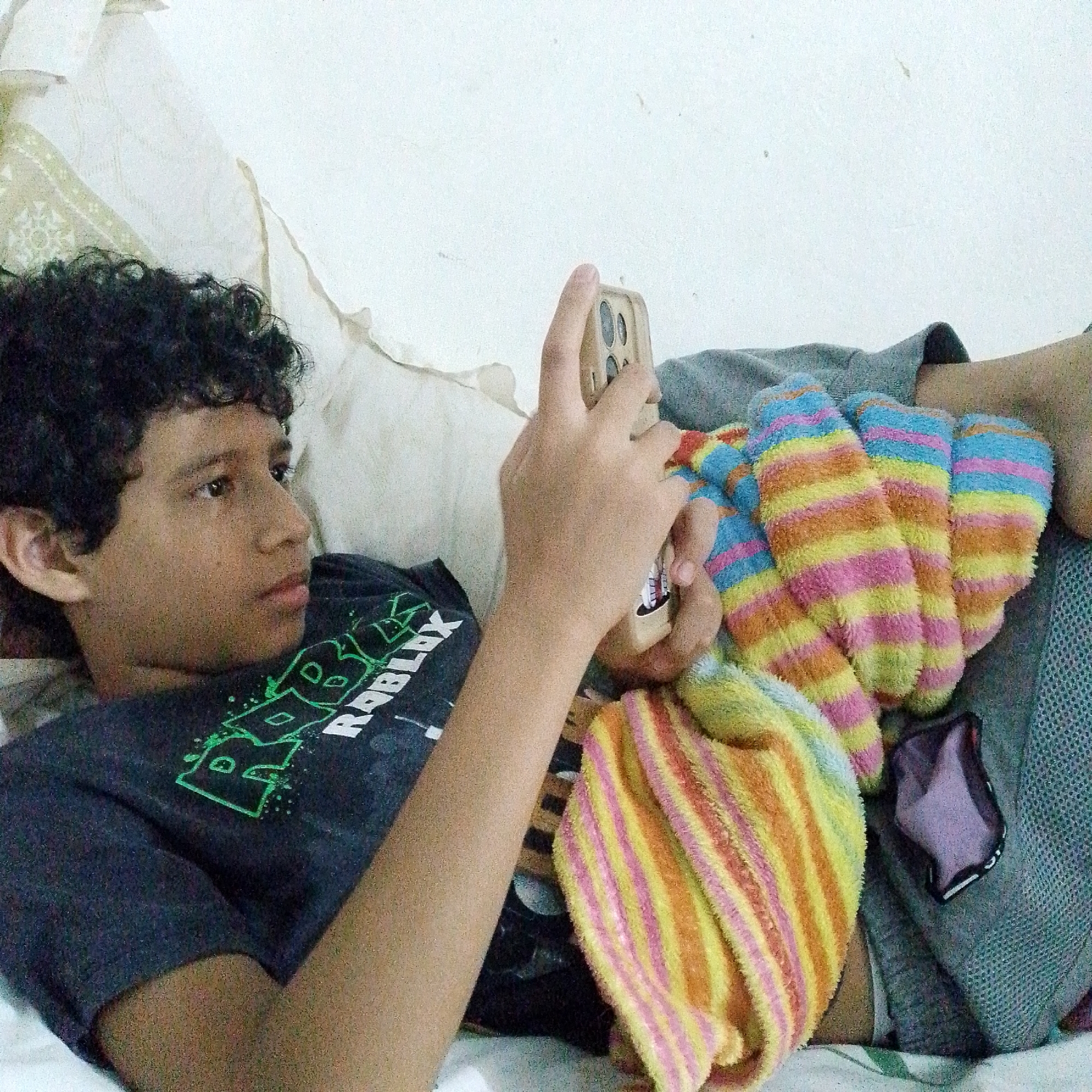 A teenager is lying on a bed, looking at a cell phone. The teenager is wearing a dark gray t-shirt with a green Roblox logo. A striped towel, mostly yellow, orange, and pink, is resting on the teenager's legs. A blue-ish grey knit fabric is also present. The teenager is in a room with white walls and is resting against a white pillow. The location is Tinaquillo, Venezuela.