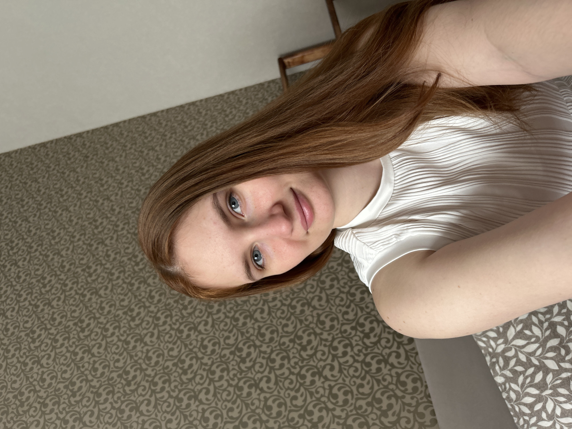 The image is a selfie of a fair-skinned person with light brown or auburn hair and blue eyes. They are wearing a white top with a textured or ribbed pattern. The background consists of a patterned carpet or flooring and a portion of a white wall. A wooden chair is partially visible in the upper right corner. The image does not provide enough details to confirm or deny if this image was taken in the city of Chaikovsky, Russia.