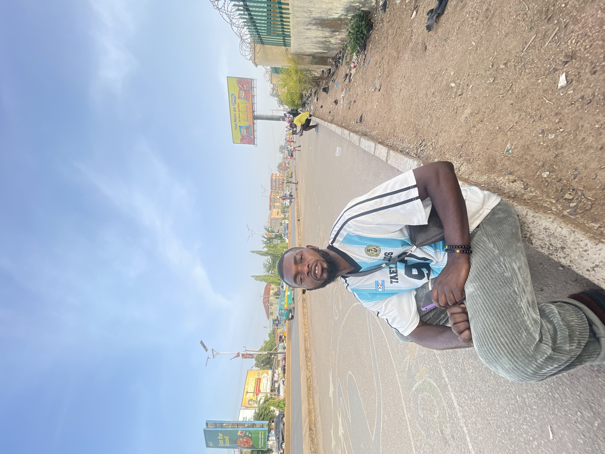 This outdoor image, taken in Jos, Nigeria, features a dark-skinned man with a beard, smiling at the camera while kneeling on the side of an asphalt road. He is wearing a light blue and white striped jersey, reminiscent of an Argentina football kit, with "TAEZLUS" and the number "6" printed on it, along with a crest. He also wears grey corduroy pants and a black beaded bracelet on his right wrist, holding a purple phone in his left hand. A black shoulder bag is visible on his left side.
The road surface has faded white markings, possibly from a children's game. Adjacent to the road is a dry, dusty dirt path strewn with debris and small rocks. A low concrete wall topped with razor wire runs along this path, with sparse green vegetation visible beyond it.
In the background, the road extends into the distance under a clear, bright blue sky, indicating a sunny day. Several other people are visible along the roadside, including one person in a yellow shirt crouching further down. Various vehicles, including cars and possibly a bus, can be seen on the road. Buildings line the street in the distance. Modern street light poles are also present.
Visible text includes "TAEZLUS" and "6" on the man's jersey. A yellow billboard in the mid-ground reads "You Have an ONGA," likely for ONGA seasoning. Another blue billboard on the far left displays "Eat for Good" vertically, and part of a billboard closer to the center indicates "TOPCITY MARKETS."