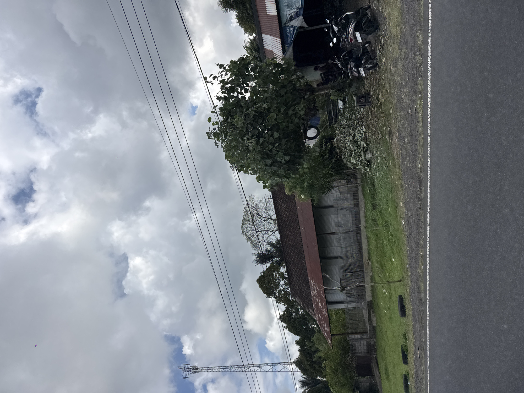 The image depicts a roadside scene in Susut, Indonesia. In the foreground, a grey asphalt road with a white line is visible. Beyond the road, a grassy embankment leads up to a weathered building with a corrugated metal roof. The building appears to be a long, single-story structure with a fence made of vertical wooden slats. To the left of the building, a tall telecommunications tower stands against a cloudy sky. A cluster of trees with dense foliage is situated to the right of the building, partially obscuring another structure with a similar corrugated metal roof, possibly a shop. Two motorcycles are parked in front of this second structure. The overall environment suggests a semi-rural or small town setting.