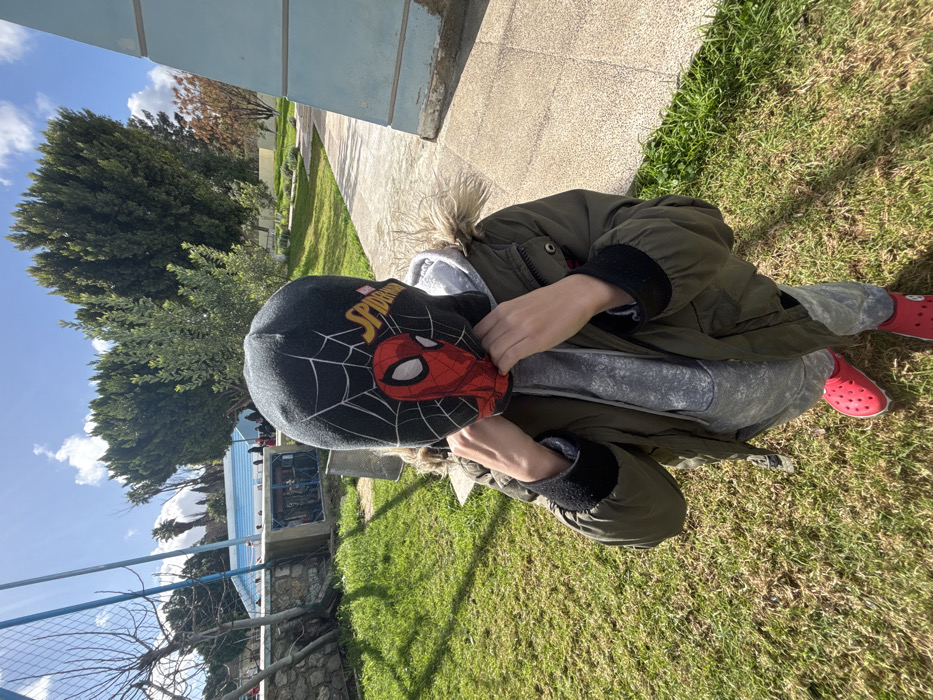 An outdoor, eye-level photograph depicts a child standing on a patch of green grass under a bright, partly cloudy sky in what appears to be a public park or schoolyard environment in Meknes, Morocco. The child, centrally positioned and facing the camera, wears a grey hooded sweatshirt beneath an olive green winter jacket with a fur-lined hood, grey pants, and red Crocs-style footwear. Their face is entirely covered by a dark grey balaclava or beanie featuring a red Spider-Man mask design and yellow "SPIDER-MAN" text. The child's hands are raised, appearing to adjust the lower edge of the balaclava around their chin and neck area.
In the midground, to the left of the child, a low, light-colored stone wall separates the foreground grass from an area containing a light blue, horizontally banded structure, possibly a building or public facility, with several indistinct human figures visible near it. Adjacent to this wall, a small, open concrete utility box with visible internal components is present, alongside a section of dark metal fencing and bare tree branches. To the right of the child, a light-colored paved pathway runs towards the background, parallel to a weathered light blue concrete pillar. The distant background is dominated by dense, tall green trees, consistent with conifers, and scattered smaller shrubs. The ground is primarily green grass with some drier patches. The scene is illuminated by natural sunlight, casting shadows.