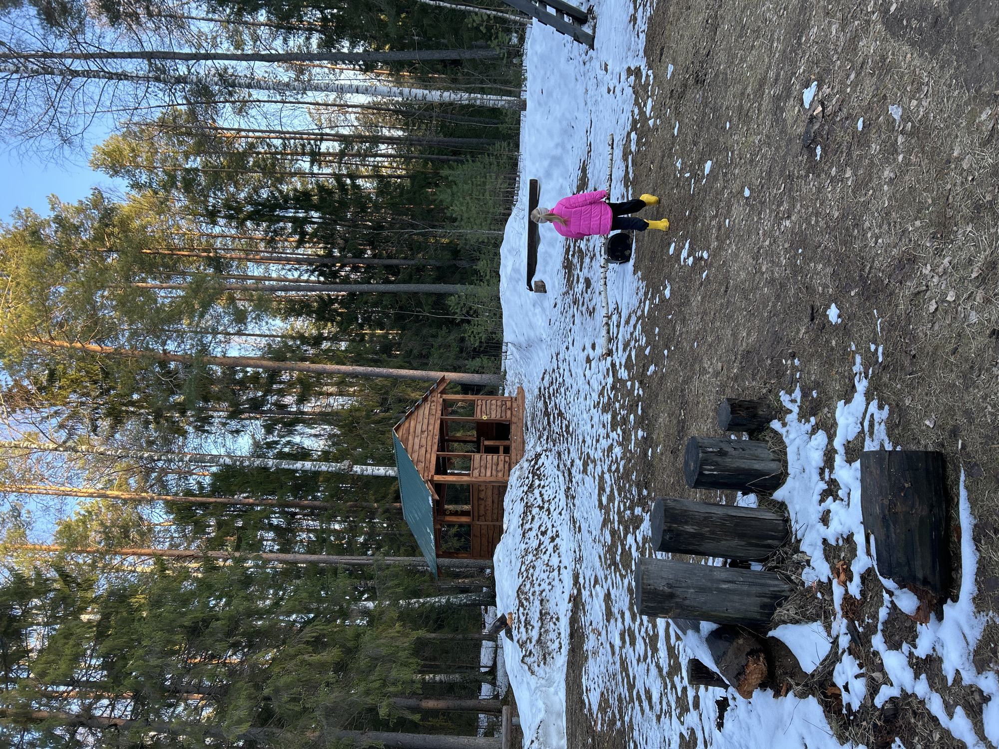 The scene depicts a partially snowy, wooded area. On the left, tall trees reach upwards towards a blue sky. A small wooden playhouse with a green roof stands amidst the trees. A woman in a pink jacket and yellow boots is seated on a wooden bench in the center of the frame, positioned on a patch of snow mixed with dry ground. Adjacent to her are log-style steps or seating. The environment suggests a rural or park setting, possibly located in Noviy, Russia.