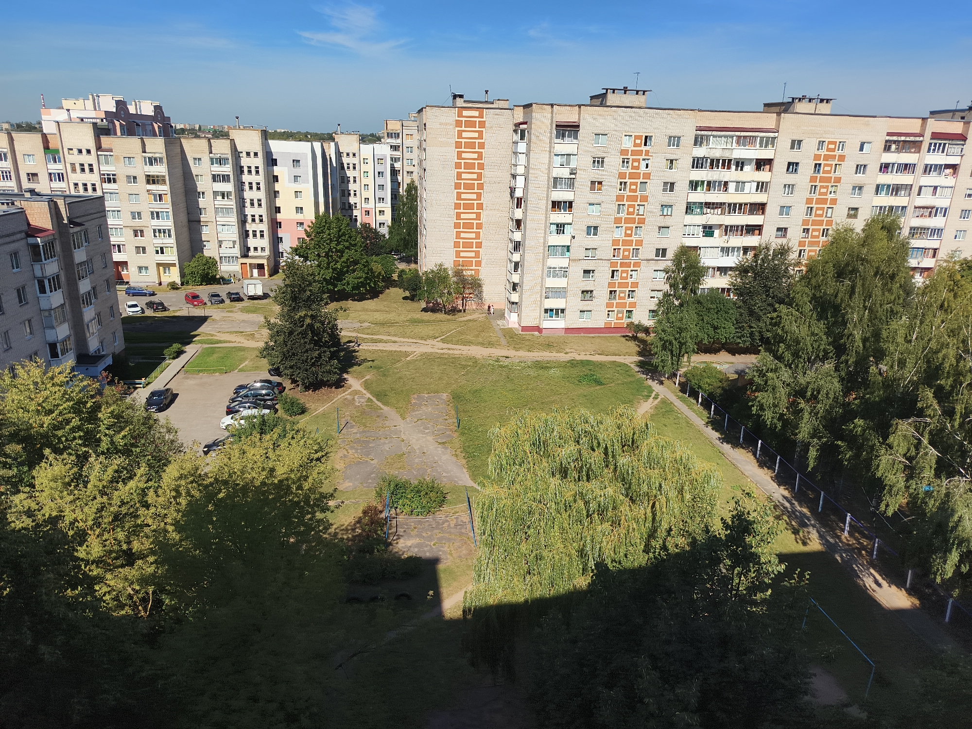 The image captures an aerial view of a residential area in Baranavichy, Belarus, on a clear, sunny day. The scene is dominated by several multi-story apartment buildings, typical of Soviet-era architecture, with light-colored facades and numerous windows. Some buildings feature decorative brickwork in orange or beige patterns. In front of the buildings, a patch of green grass is interspersed with concrete pathways and a small, paved area that appears to be a former courtyard or playground. A cluster of parked cars is visible on one of the concrete surfaces, including a white car, a red car, and several darker-colored vehicles. Trees, some with lush green foliage and others with a weeping willow-like appearance, are scattered throughout the open spaces between the buildings and along the edges of the scene, casting shadows on the ground. A volleyball net and posts are set up in one of the open grassy areas. The overall impression is one of a typical urban residential environment during daylight hours.