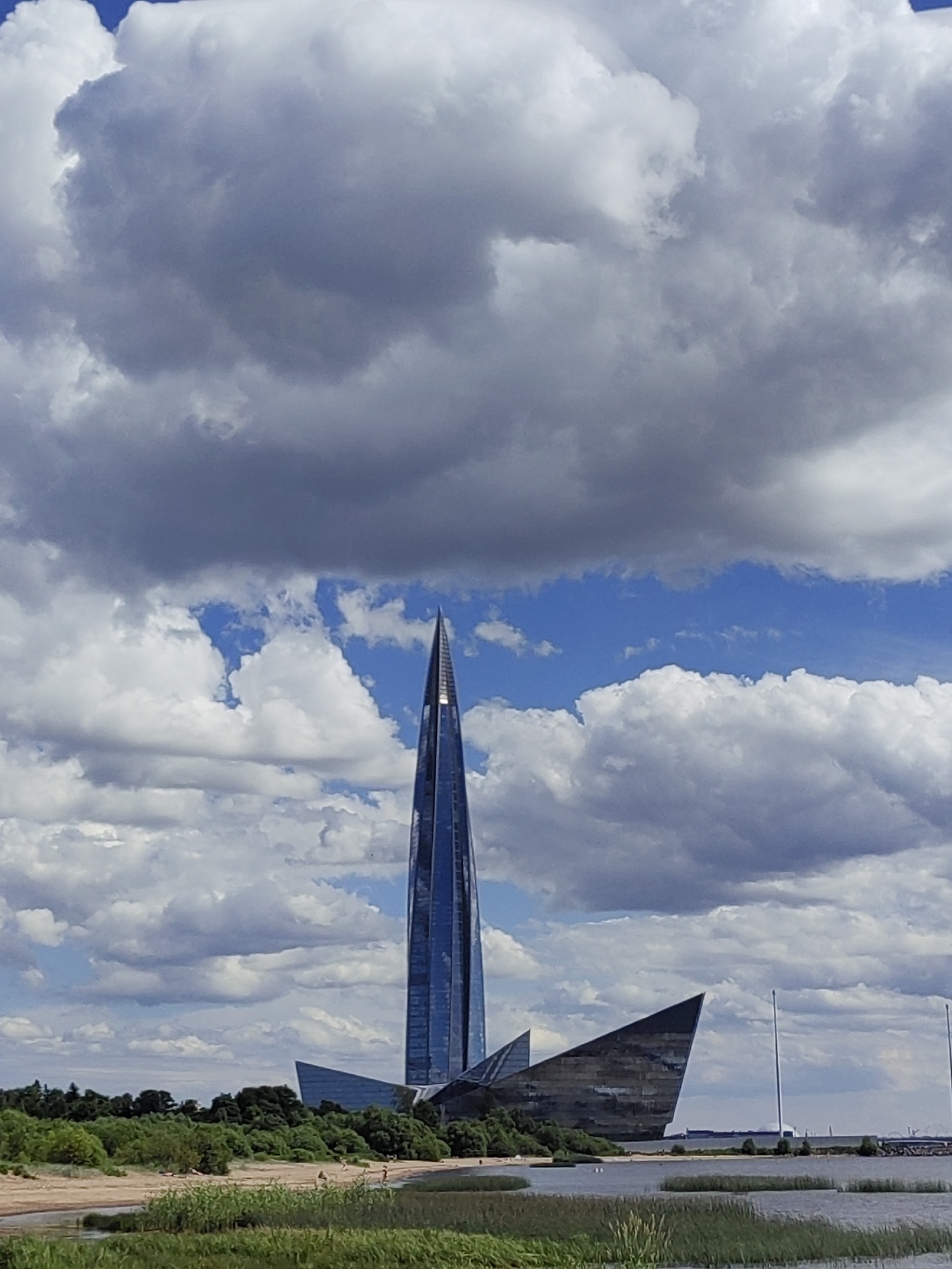 The image shows the Lakhta Center, a skyscraper in Saint Petersburg, Russia, on a cloudy day. The skyscraper is a tall, slender, glass-covered building with a pointed top, reflecting the blue sky and white clouds. In front of the skyscraper, there is a curved, dark-colored structure with sharp angles, also made of glass. The foreground features a body of water with tall grass growing in it, and beyond that, a sandy beach with scattered trees and vegetation. Several small figures, presumably people, are visible on the beach. The sky is filled with a dramatic mix of white, fluffy clouds and darker, more ominous clouds, suggesting a dynamic weather system. The overall scene depicts a modern architectural marvel set against a natural landscape with a hint of urban development in the distance.