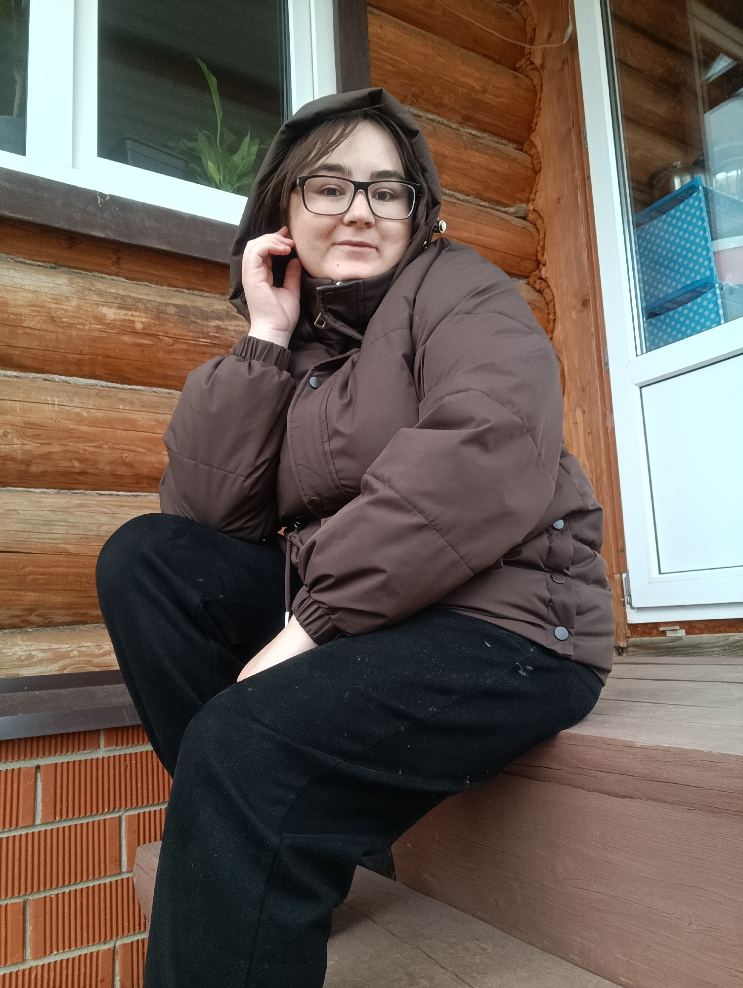 A woman with glasses, wearing a brown hooded puffer jacket and black pants, sits cross-legged on a wooden step outside a log cabin. The cabin has white-framed windows. To the left of the frame is a brick wall. The location is likely near Barda, Russia, given the cabin architecture and the presence of windows. The woman is holding her hand near her face, possibly resting or adjusting her hood.