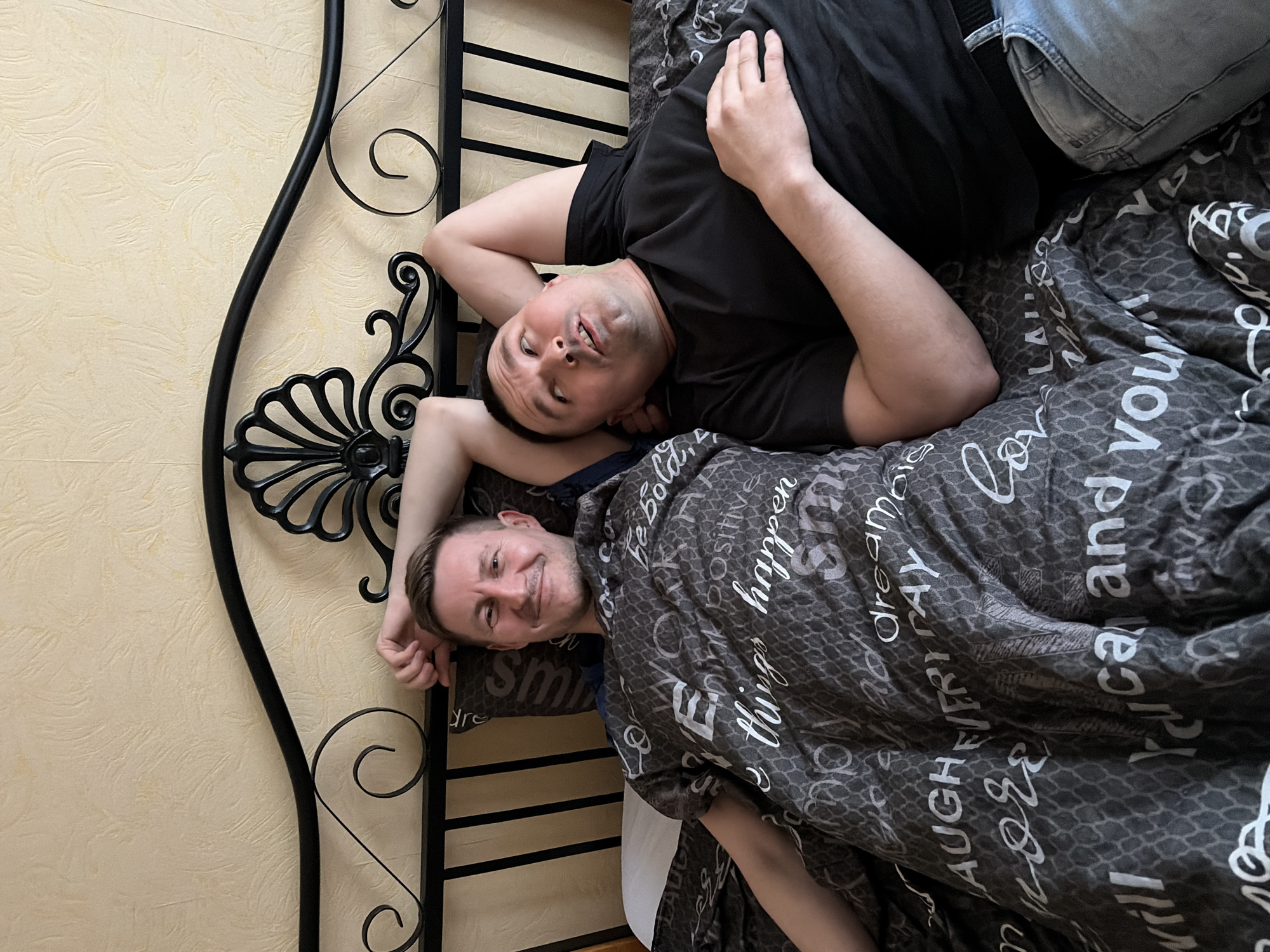Two men are lying on a bed with a black metal frame headboard. One man is lying closer to the headboard, covered with a gray blanket printed with white words. The other man is lying on his back with his arms above his head, wearing a black t-shirt and jeans. The wall behind the headboard is textured and light yellow. The photo is rotated such that the headboard is to the left. There is no information about the location in the city of Izhevsk, Russia.
