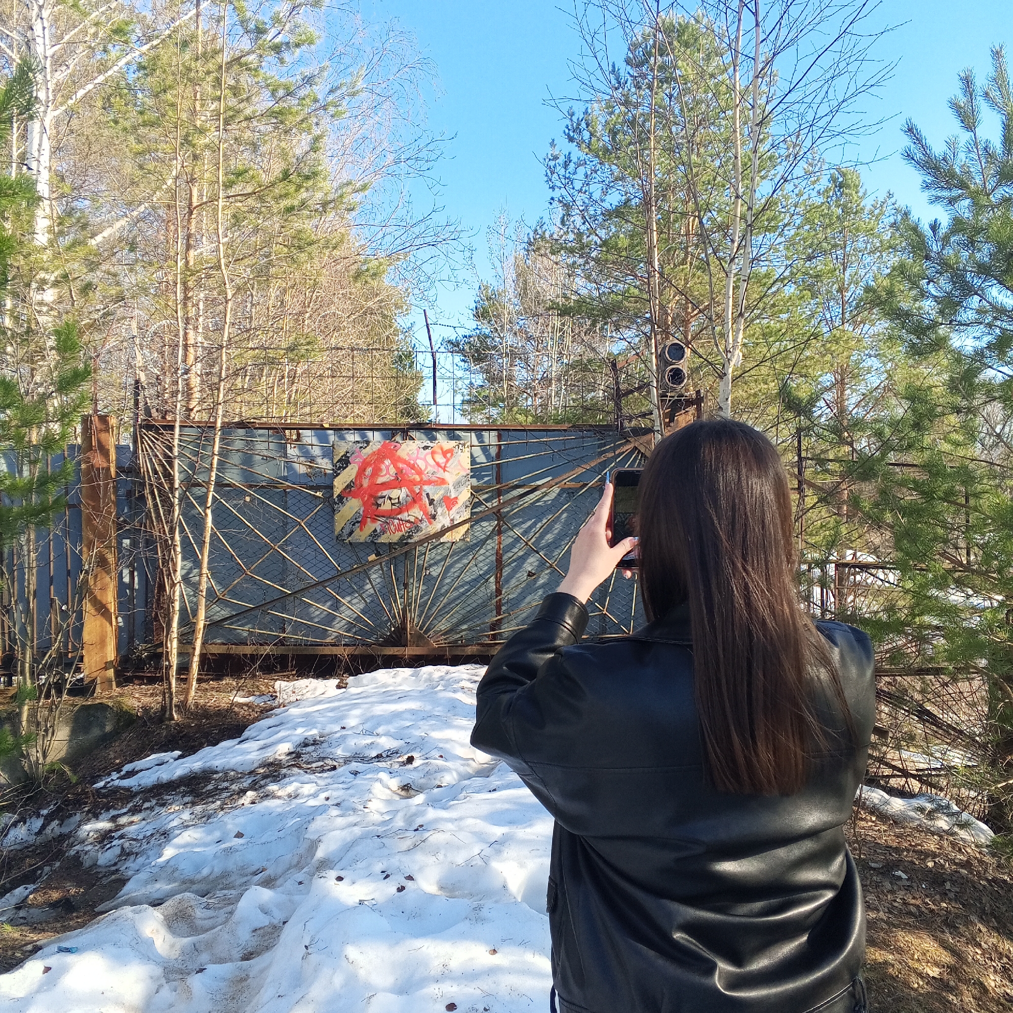 A woman with long brown hair is photographing an abandoned gate with graffiti on it. She is wearing a black leather jacket and holding a smartphone in her hands, pointing it towards the gate. The gate is grey and covered in rust, with a square section in the middle containing a red anarchy symbol and other markings. The gate is partially obscured by trees. In the foreground, there is a snow-covered area with patches of dirt visible. The scene is set in a wooded area with tall trees and a clear blue sky. The location is Chaikovsky, Russia.