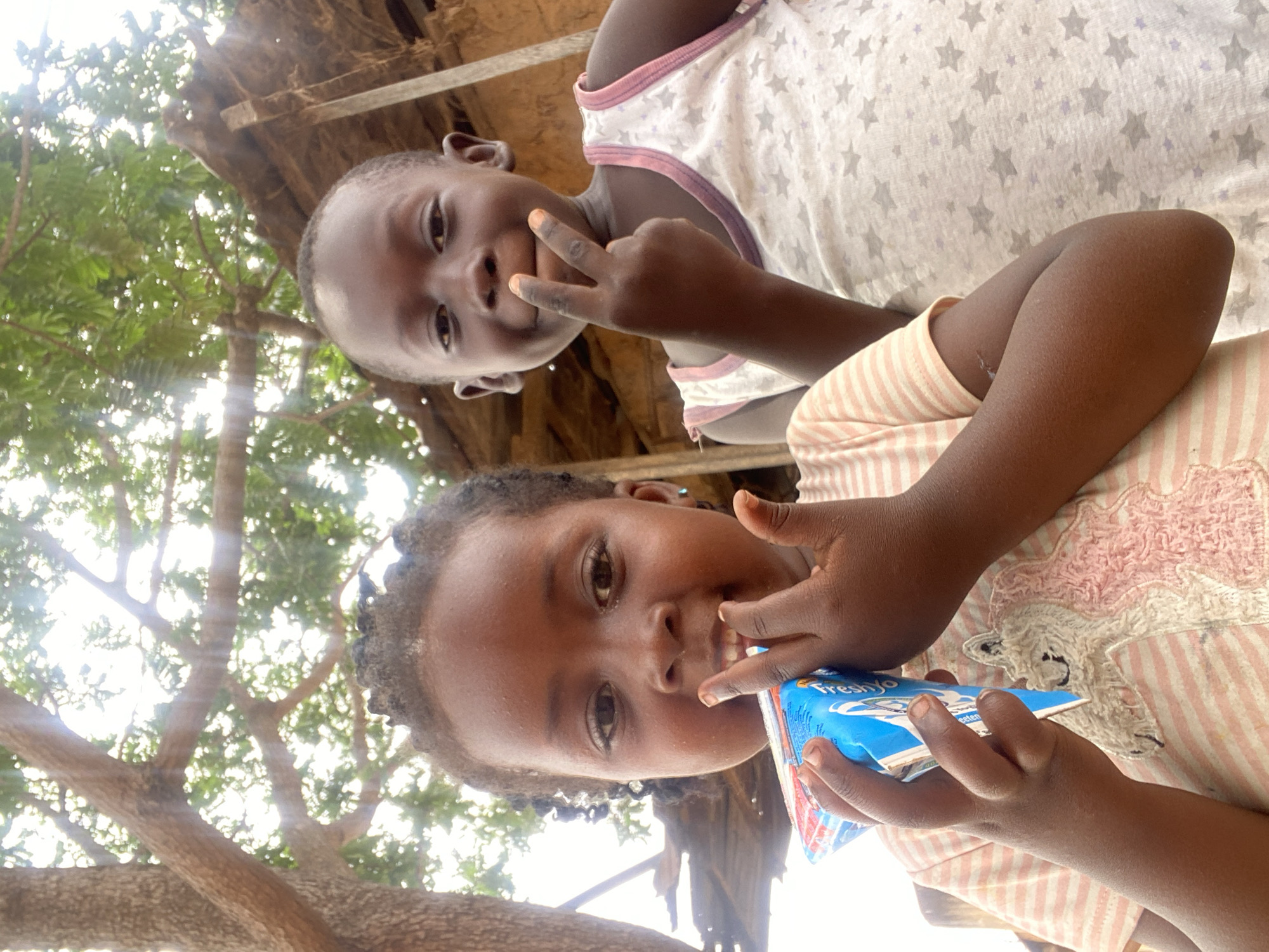 Two young girls are pictured from a low angle in what appears to be Kuru Dungus, Nigeria. The girl closer to the viewer holds a blue and white juice box labeled "Fresh Yo." She is wearing a pink and white striped top and has curly dark hair. The second girl is in the background, wearing a white tank top with star patterns. She is holding up two fingers in a peace sign. The background features tree branches and glimpses of what seems to be a wooden structure. The setting suggests an outdoor environment in Kuru Dungus.