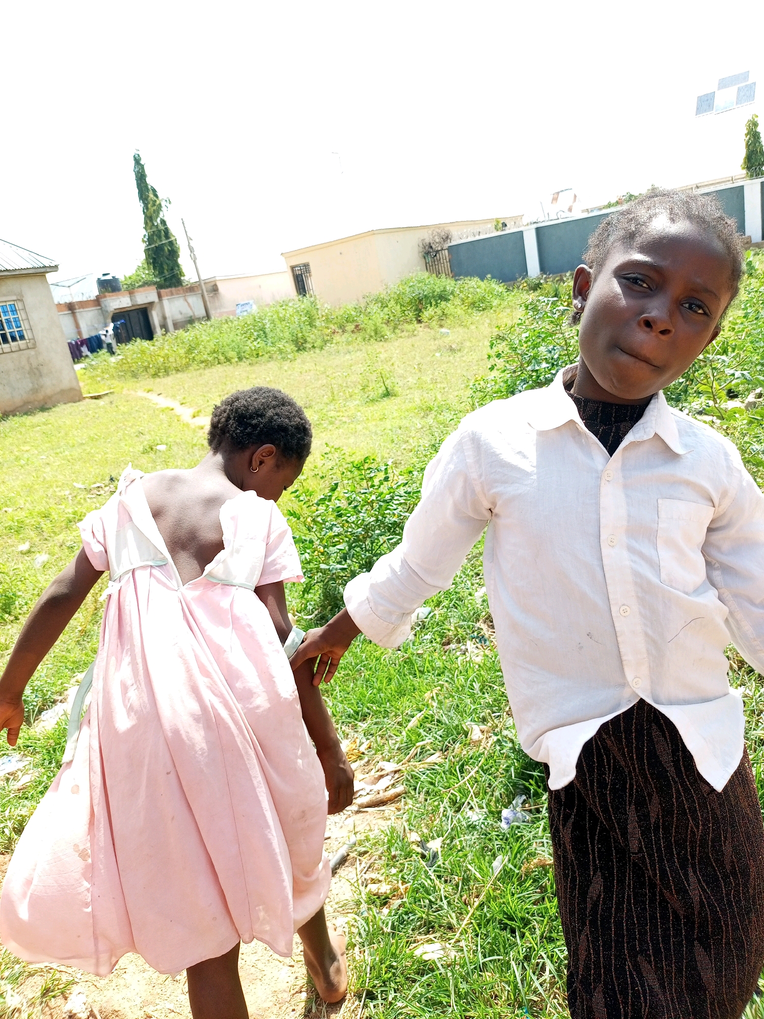 In Tempure, Nigeria, two young girls are captured in an outdoor scene. The girl in the foreground, facing the camera, wears a white button-down shirt over a black, patterned garment, her expression curious. To her left, a second girl, with her back to the camera, is dressed in a light pink dress. The environment appears to be a grassy area with some dry debris, likely a yard or an open space within a residential area, indicated by a building with a blue-trimmed window and a water tank visible in the background. The sky is bright and overcast. The girls are interacting, with the girl in the foreground holding the hand of the girl in the pink dress.