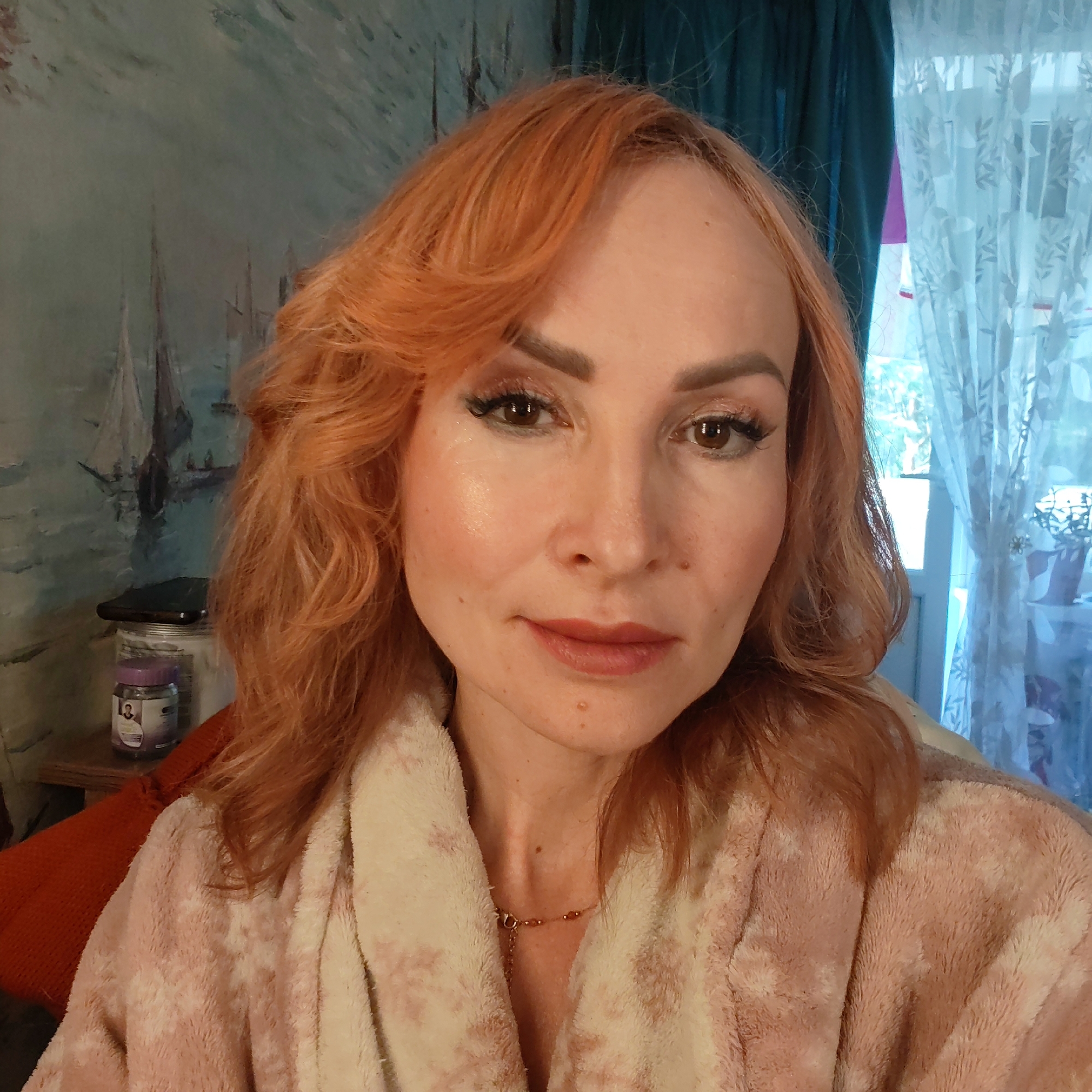 This indoor portrait, set in Chaikovsky, Russia, features a woman with auburn, wavy hair and brown eyes, captured in a selfie-style close-up. She is wearing a soft, light pink and beige patterned fluffy bathrobe, with a delicate gold-colored necklace adorned with small beads visible around her neck. Her face shows light makeup, including eyeshadow, eyeliner, mascara, and a natural-toned lipstick. Her expression is calm and composed, with a subtle, gentle smile.
To her left, a large painting or wall mural depicts sailboats on a muted blue-green body of water, suggesting a maritime or lakeside scene. Below this on a shelf, a dark-colored smartphone, a large white jar, and a smaller purple jar with indistinct text are visible.
To her right, a window allows natural daylight to enter, covered by sheer, light-colored curtains featuring a leaf pattern, layered over darker blue-green curtains. A potted plant can be vaguely seen through the window. A small portion of orange, textured fabric, possibly a cushion or blanket, is visible in the bottom left corner of the frame. The overall setting suggests a comfortable, homely environment during the daytime.