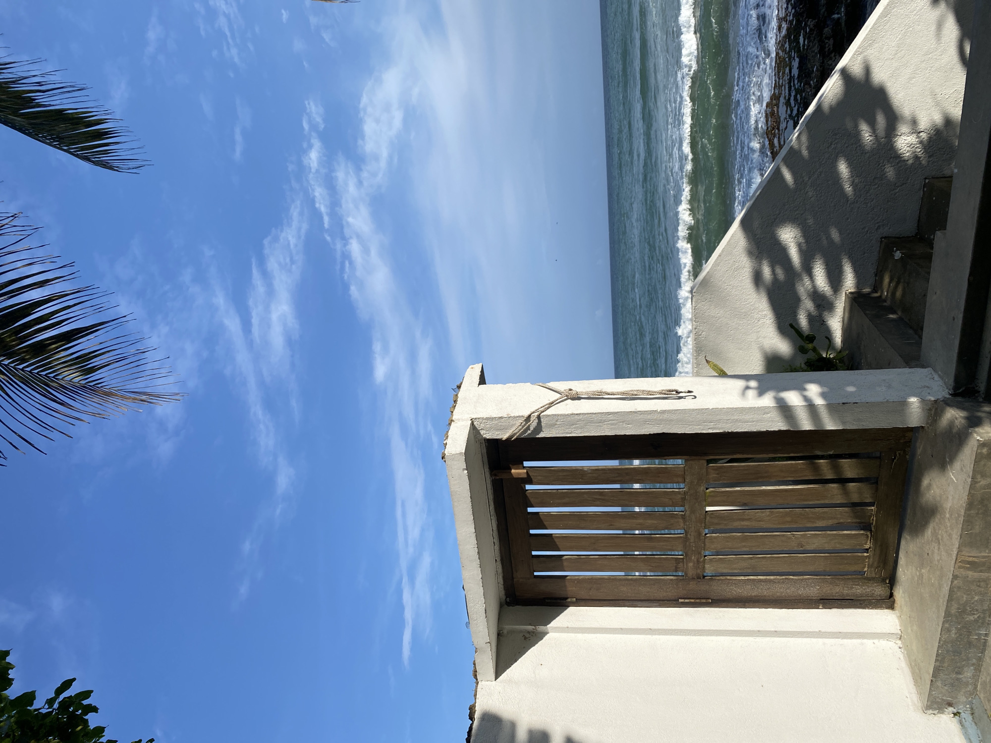The image depicts a partial view of a coastal scene in Goviyapana, Sri Lanka. In the foreground, a weathered, light-colored concrete structure encloses a wooden slatted gate. A worn rope is tied near the top of the concrete frame. The sea, showing waves, abuts the right side of the structure. To the left, the sky is blue with wispy clouds. Palm fronds are visible in the upper left corner, and some foliage is visible in the lower left. Shadows are cast on the concrete steps to the right of the gate, indicating sunlight.
