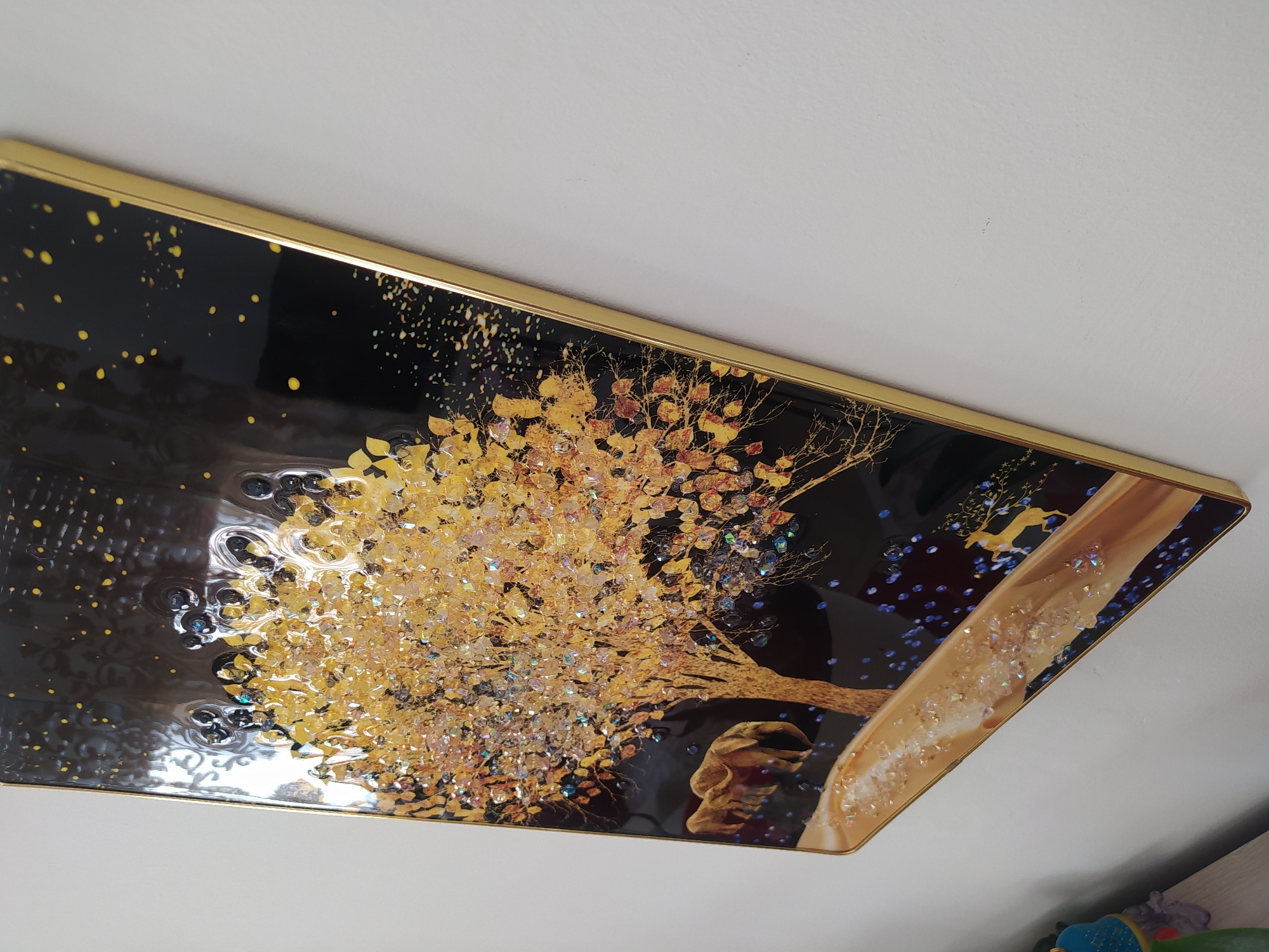 The image shows a framed piece of art hanging on a white wall. The artwork features a golden tree with crystalline leaves against a black background. There are also two golden elephants depicted in the lower left portion of the artwork, and a path with embedded crystals in the foreground. The frame is thin and gold-colored. The image is taken at an angle, showing the side of the frame and the wall. There is no indication of the city of Livorno, Italy, in the image.
