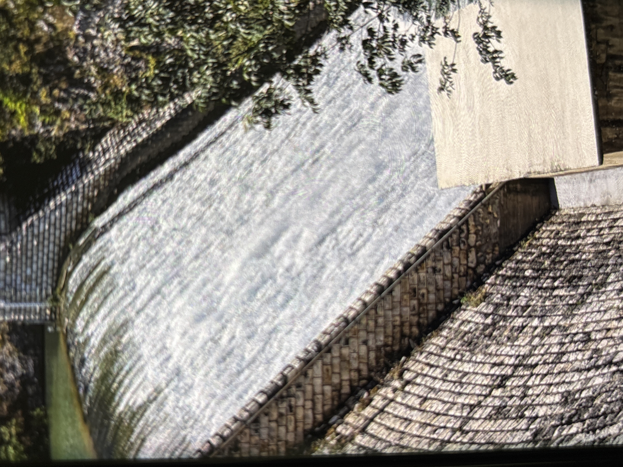 The image presents an aerial or elevated view of a dam or weir, where water cascades vigorously over a man-made structure under bright daylight.
In the central area, a wide expanse of water flows over the barrier, creating a dynamic, shimmering surface with numerous ripples and reflections, indicating rapid movement and direct sunlight. Dense green foliage, likely from trees or bushes, occupies the upper left and left edges of the frame, partially extending over the water and casting shadows.
To the right of the flowing water, a textured stone embankment or wall rises, featuring a patterned surface that suggests brickwork or stone tiling. A dark railing or fence runs along the upper edge of this embankment. Further towards the upper right, a large, light-colored, smooth concrete or stone slab is visible, potentially a platform or part of an adjacent structure. Below this slab and next to the embankment, another textured, possibly sloping, surface is visible, resembling a stone-tiled roof or cobbled area.
The perspective is a downward, diagonal shot, with the water appearing to flow from the upper left towards the lower right of the image. The scene conveys a sunny day, with strong highlights on the water and defined shadows from the surrounding foliage. There are no people or visible text. The combination of water control structures and natural elements is consistent with river or canal systems found in areas like Wolverhampton, United Kingdom.