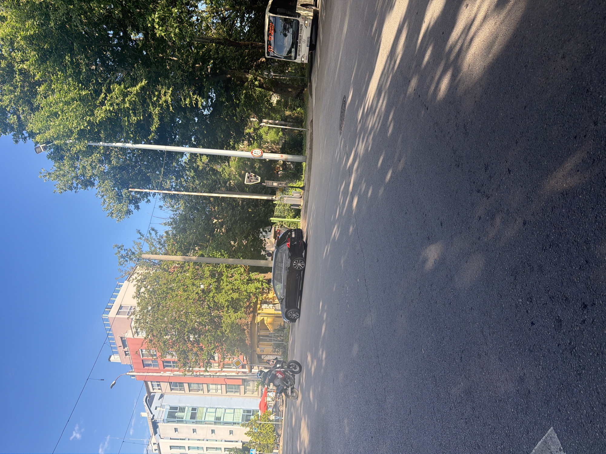The image shows a street scene in Plovdiv, Bulgaria. The road occupies a significant portion of the frame, with a black car parked on the side of the road. A motorcycle is parked adjacent to the car. Buildings of varying colors and heights line the opposite side of the street. Trees are present on both sides of the street. A bus is visible near the top of the frame. The sky is blue and clear. Tree shadows are cast on the street. A street light is visible. A speed limit sign is visible.
