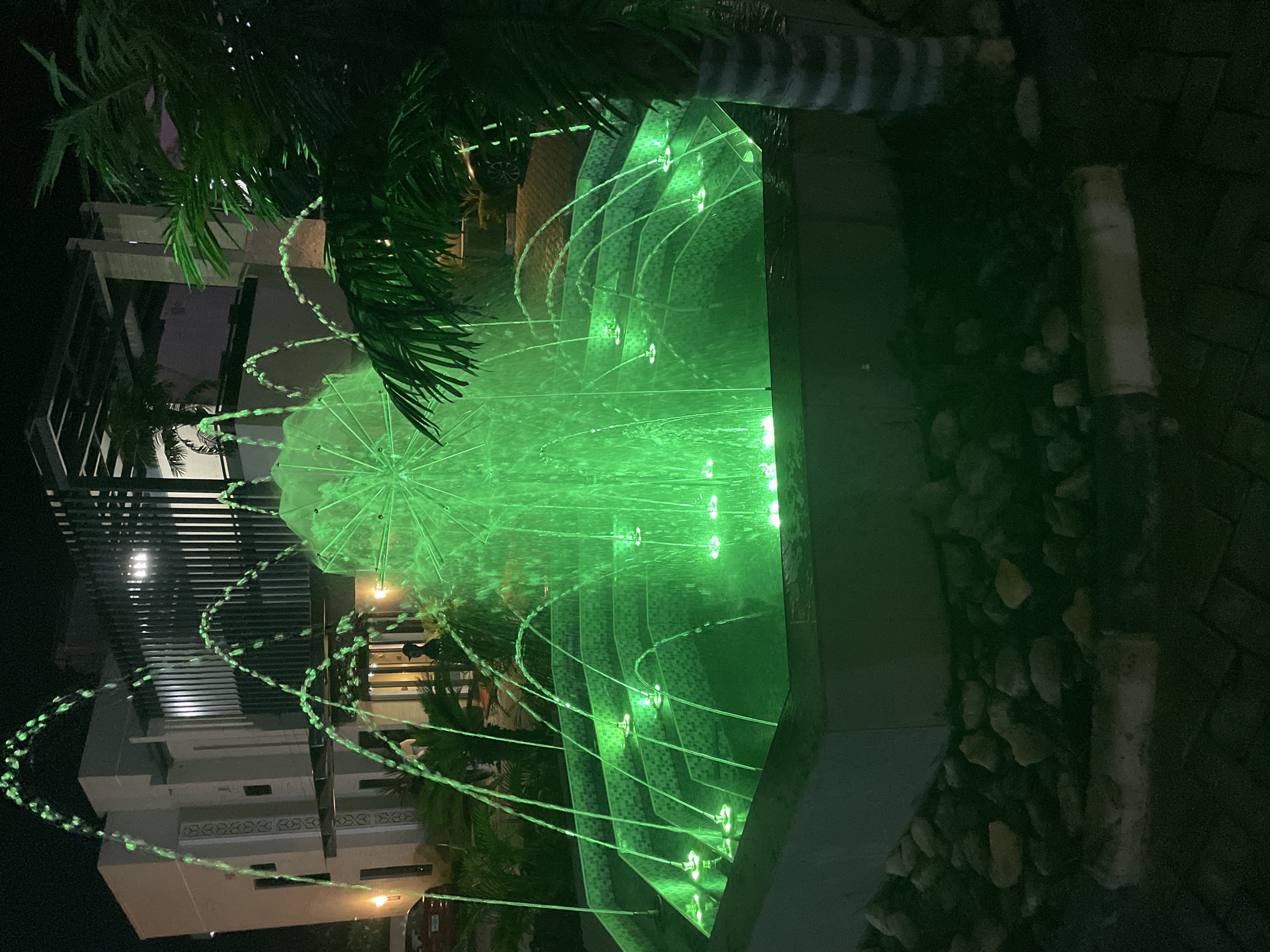 This image shows an illuminated fountain with green lights at night. The fountain is in the foreground, with water jets arcing upwards. Behind the fountain, there is a modern building with lights on. Palm trees are visible on the right side of the image. The scene appears to be an outdoor space, possibly a courtyard or garden, attached to the building. The image was taken in Akwanga, Nigeria.