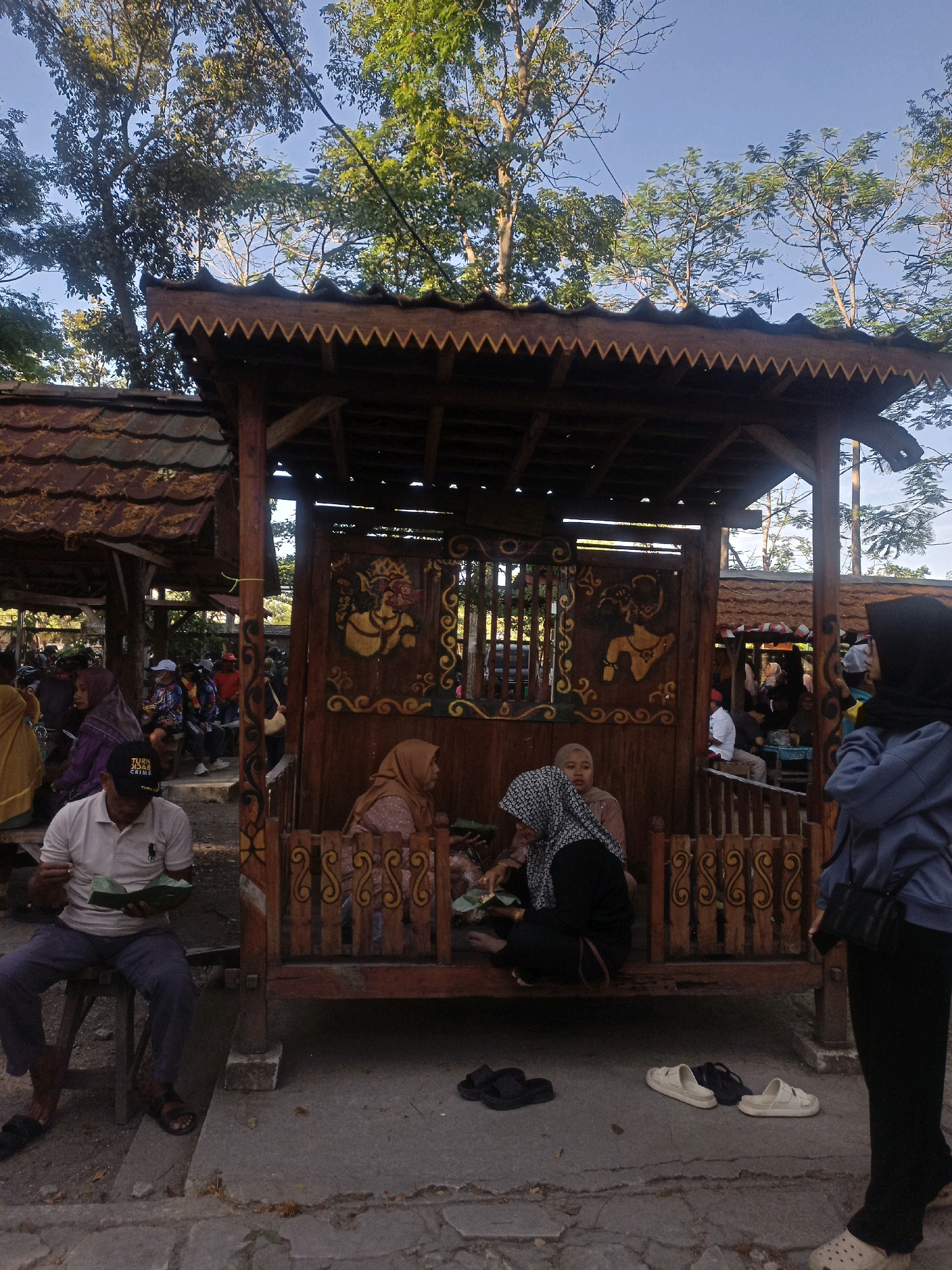 In the city of Karungan, Indonesia, at what appears to be an outdoor eating area, several individuals are present. The scene is dominated by a wooden structure with a decorative, jagged-edged roof. Inside this structure, three people are seated, two women wearing headscarves, and a third person partially obscured. One woman is holding a small green object, possibly food wrapped in leaves.
In the foreground, a man is seated on a stool to the left, also holding a green wrapped object. He is wearing a white polo shirt and dark pants, with sandals on his feet. To the right, another person, wearing a blue jacket and black pants, stands facing away from the camera. Various footwear, including sandals and slippers, are scattered on the ground.
The background reveals more people seated at tables, suggesting a bustling atmosphere. Trees with green foliage and a clear blue sky are visible above the wooden structures. The ground is paved with rough stone. The overall impression is of a casual, communal gathering or dining experience in an outdoor setting.