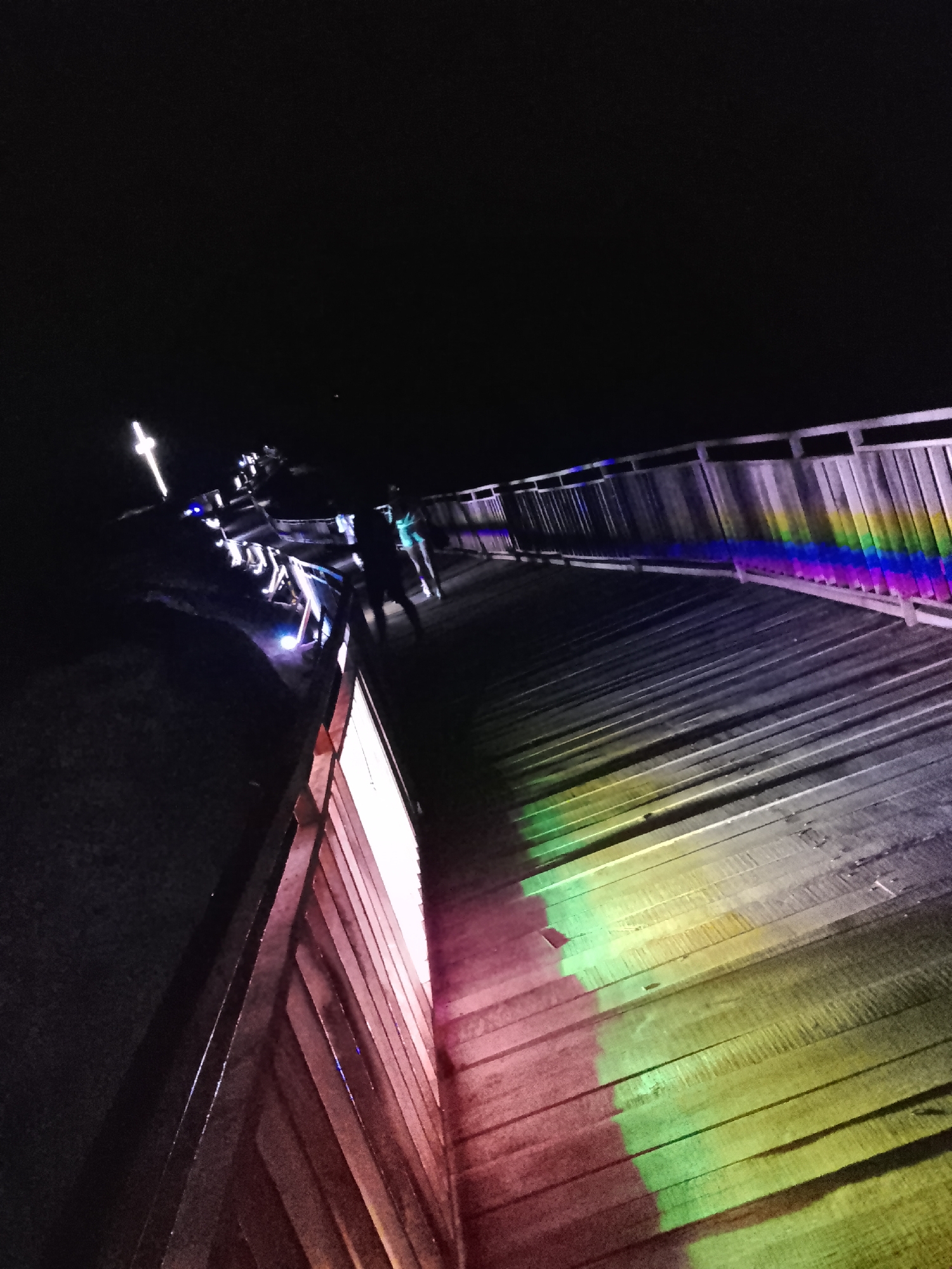 This image depicts a wooden walkway at night, illuminated by various colored lights, likely part of an art installation or event. The walkway is constructed of horizontal wooden planks and features a railing along one side. Prominent rainbow-colored light reflections are cast onto the walkway and railing, suggesting a light source projecting these colors. Several individuals are visible on the walkway, appearing to be walking or standing. The overall environment is dark, indicating it is nighttime. The location is identified as being in the city of Provadia, Bulgaria, specifically at the Astrea Observatory, which features illuminated walkways and artistic installations.