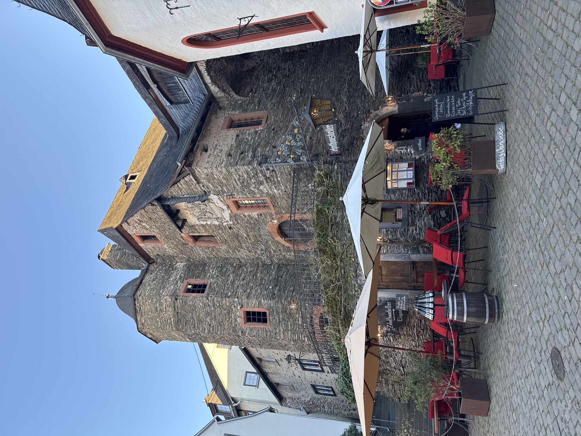 The image captures a street-level view in Beilstein, Germany, featuring the Zehnthaus and its adjacent outdoor dining area. The Zehnthaus, a historic stone building, dominates the left side of the frame. It has multiple stories, distinct windows framed in red, and a pointed turret with a spire. A sign with the name "Sehnthauskeller" is visible.
The right side of the image reveals an outdoor restaurant setting. Red chairs surround small tables under white umbrellas. Signboards are placed near the entrance, displaying a menu in German. A wine barrel is used as a table. The ground is covered with cobblestones, adding to the old-world charm of the location. The sky is clear and blue.