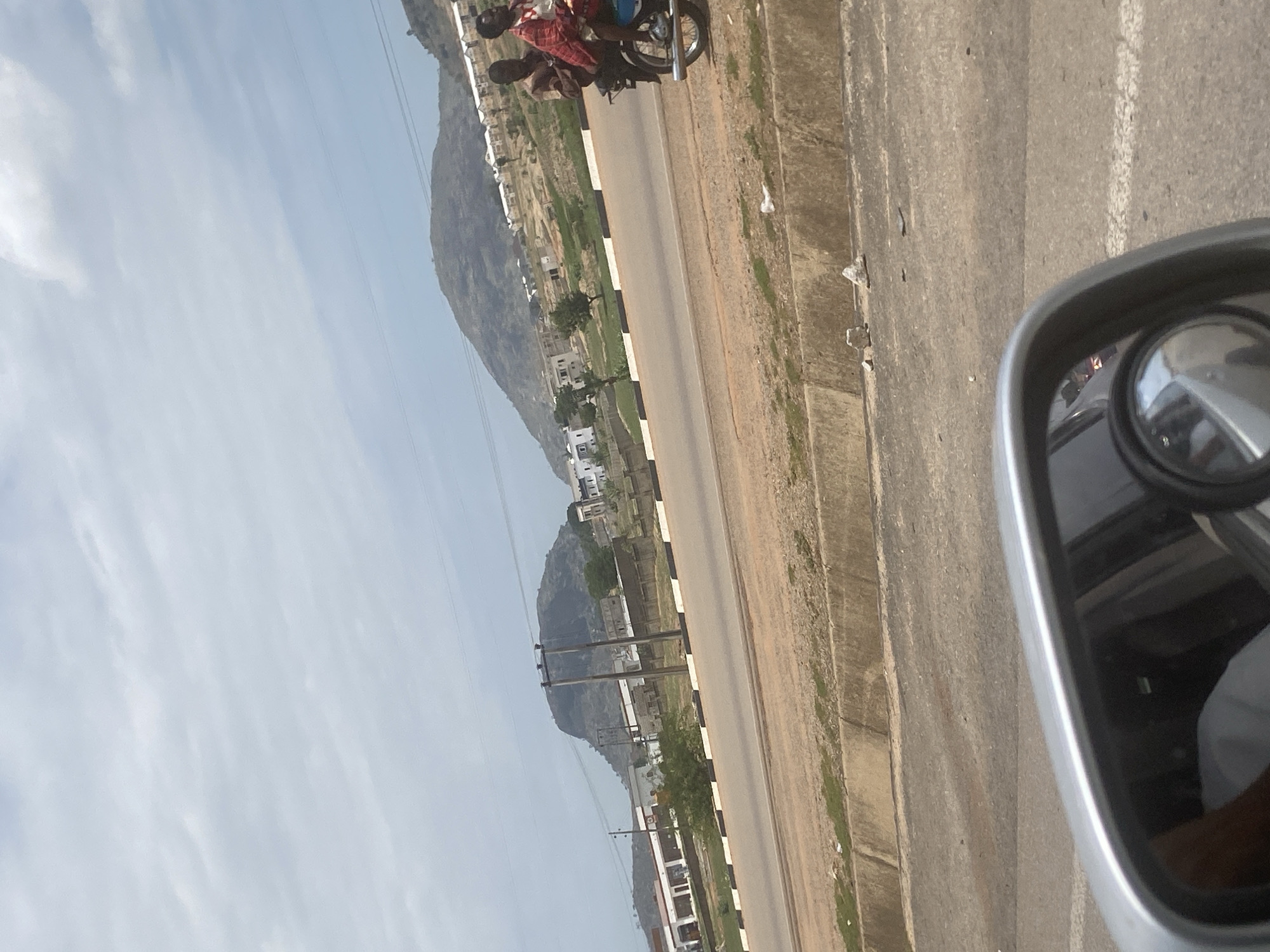 The image shows a view from a moving vehicle, possibly a car or taxi, traveling on a road in what appears to be a suburban or semi-urban area near Abuja, Nigeria. The road has a paved surface and features black and white edge markings. Along the side of the road, there is a strip of vegetation and a concrete curb. In the distance, hills or small mountains are visible, with some buildings scattered on their lower slopes. Closer to the road, there are more buildings, including what appears to be residential houses. In the foreground, the side mirror of the vehicle is visible, reflecting a blurry image of the interior. On the road, a motorcycle with at least one person on it can be seen moving in the same direction as the photographer. The sky is partly cloudy.