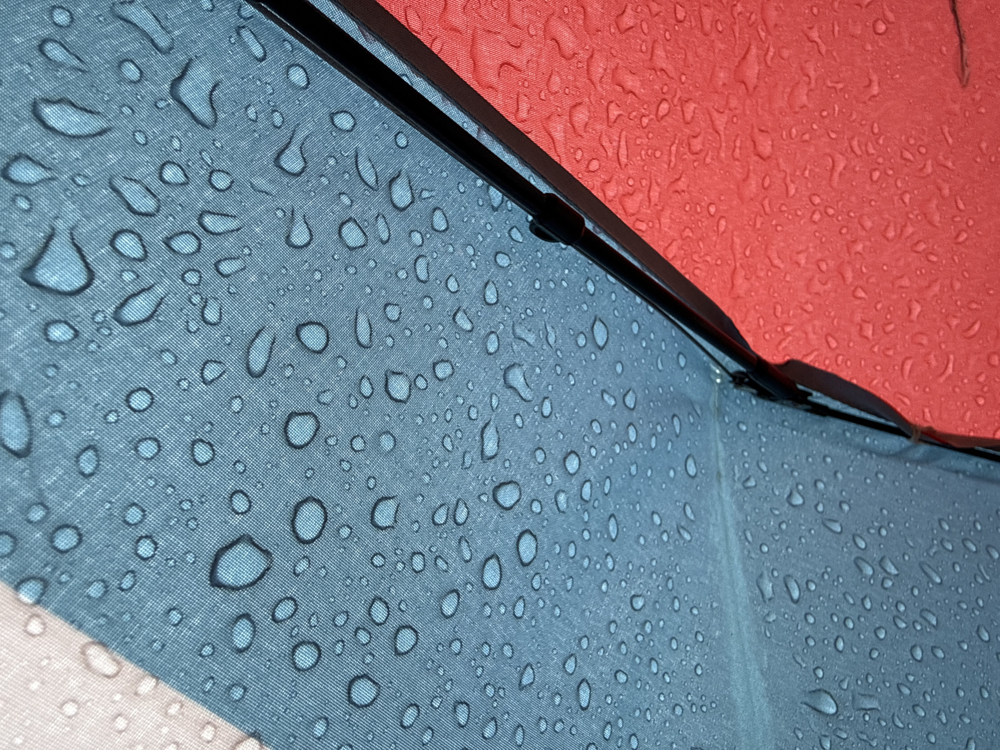 The image shows a close-up view of a multicolored umbrella covered in raindrops. The visible colors are blue, red, and white. The umbrella's fabric has a woven texture. A black support strut runs diagonally across the frame. The scene does not contain any information that would indicate the location of the city of Dmitrov, Russia.
