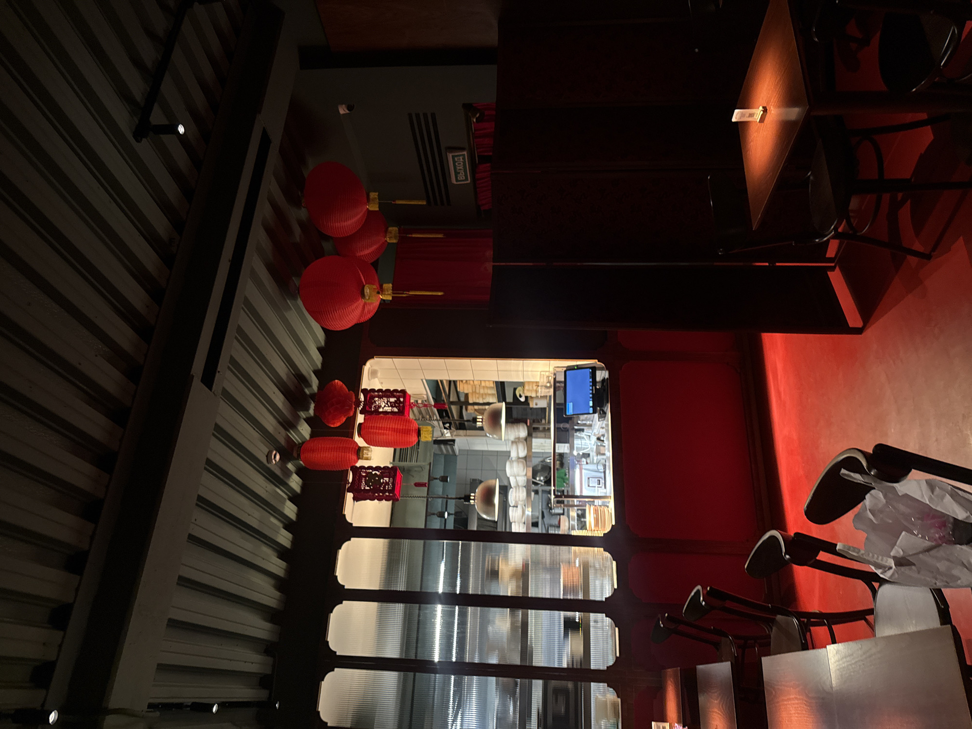 This media file captures an indoor scene, likely a restaurant or bar in Moscow, Russia, characterized by dim lighting and a strong red color scheme, possibly suggesting an Asian or themed establishment. The image is oriented sideways, presenting a horizontal view when corrected.
On the left, a dark, corrugated metal ceiling or wall runs diagonally, featuring small recessed spotlights. Below this, several red, pleated lanterns of various shapes (spherical and elongated) with gold accents are prominently displayed, hanging from the ceiling. Adjacent to these lanterns are additional ornate, dark red decorative hangings.
Towards the center, an opening reveals a brightly lit kitchen or service area with stainless steel surfaces, white tiled walls, and various commercial kitchen equipment, including what appears to be a digital screen or monitor. Below this opening, translucent, ribbed panels form a partition or window feature.
The right side of the image features dark wooden tables, some with small, illuminated objects on their surfaces, casting a warm glow. Nearby, several dark-framed chairs with light-colored seats are visible, some appearing stacked or pushed against the tables. A white bag rests on one of the chairs. The walls in this section are bathed in a deep red light, contributing significantly to the ambiance.
No people are visible in the image, suggesting the establishment might be closed or empty at the time. A white CCTV camera is visible near the ceiling on the left. A notable detail is a green sign with Cyrillic text, "ВЫХОД", clearly visible on a dark wall, indicating an "Exit". The overall atmosphere is intimate and dramatic, likely captured during the evening or night.
