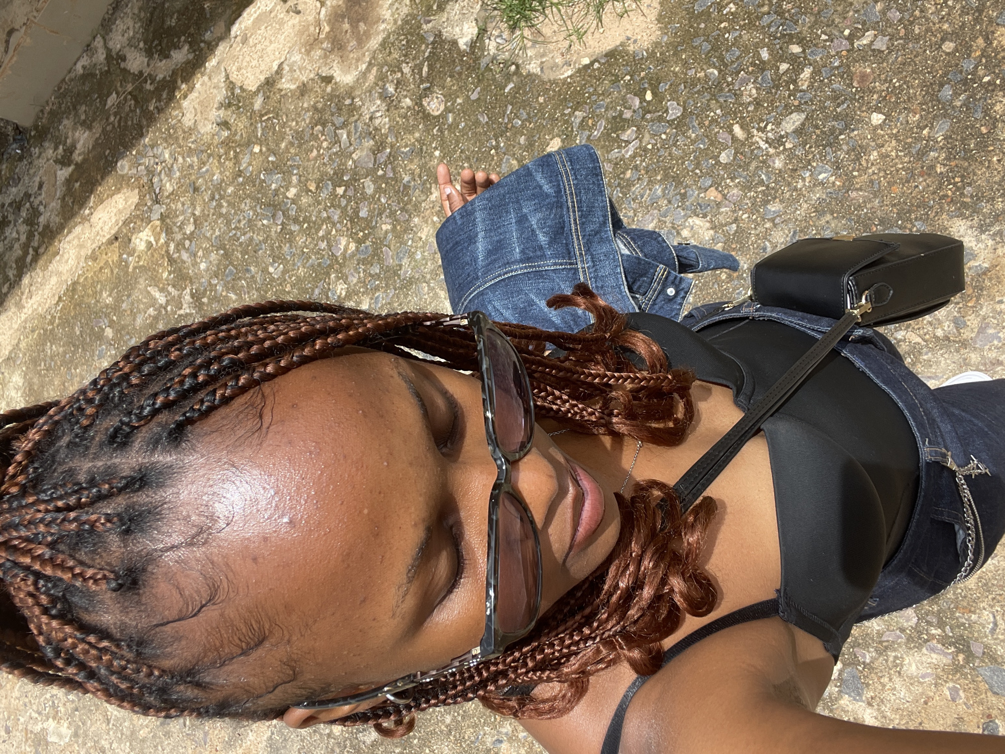 The image, captured from a high-angle perspective, shows a young Black woman standing outdoors on a surface composed of concrete and gravel. She is looking downwards, her eyes closed, and wearing dark sunglasses. Her hair is styled in numerous thin braids, with some individual strands framing her forehead. She is dressed in a black camisole top and denim jeans, accessorized with a black handbag slung across her body. A denim jacket is draped over her left arm. The background consists of an outdoor environment with a concrete structure and some vegetation visible on the right side. The lighting suggests it is daytime, with sunlight casting shadows. There are no specific identifiable landmarks that would pinpoint the location to Rafin Par, Nigeria, or any other specific city.
