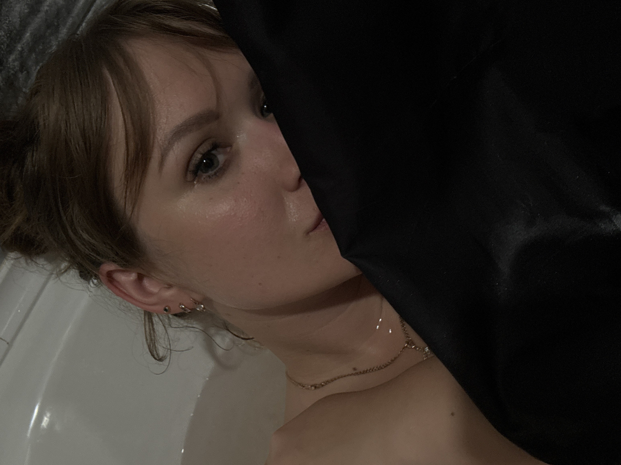 The image presents a partial view of a person, presumably female, in what appears to be a bathroom setting. The subject's face is partially visible, with light skin, brown hair styled with bangs, and visible ear piercings. Part of the subject's neck and shoulder is seen, adorned with a gold-colored necklace. A dark, possibly black fabric obscures a significant portion of the right side of the frame. The person is positioned inside a white bathtub. The location of the image is not identifiable as Chaikovsky, Russia, based on visual information.