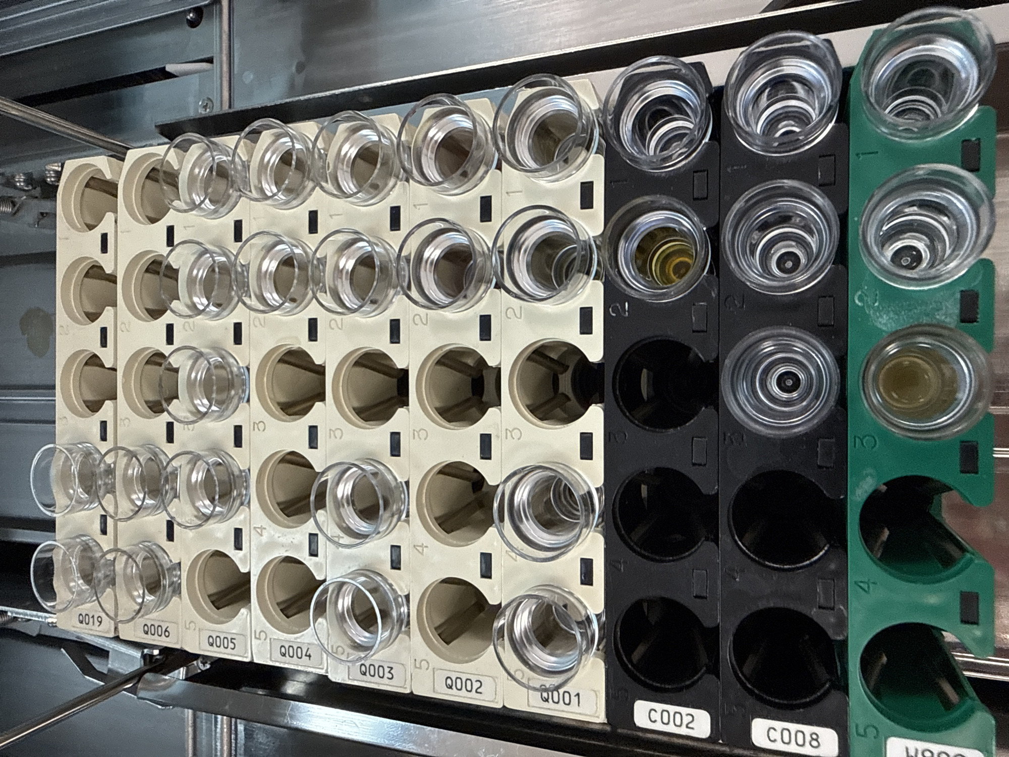 The image shows a laboratory apparatus with multiple cylindrical sample holders arranged in rows and columns. The holders are attached to three different colored racks: beige, black, and green. Each sample holder is labeled with a unique identifier, such as "Q019," "C002," and "H900." Some of the holders appear empty, while others contain liquids of varying colors and textures. The scene appears to be inside a laboratory, likely for scientific research or analysis. The location in Moscow, Russia, is not indicated by the image itself.