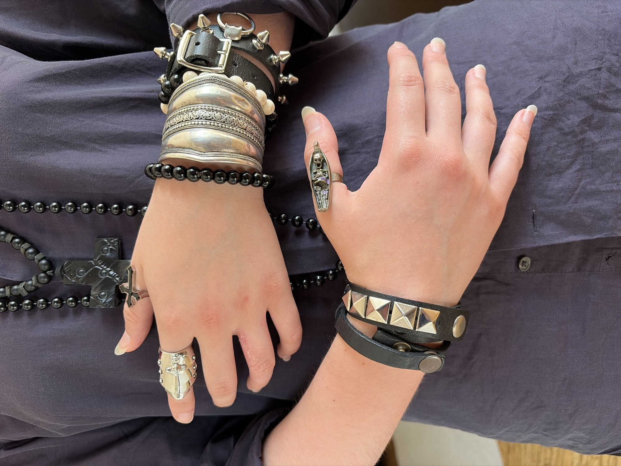 A close-up, chest-up view of a person wearing a dark purple, long-sleeved button-up shirt and multiple pieces of gothic-style jewelry. The person's hands are prominently displayed, adorned with various rings and bracelets. On the left hand, there is a large, silver, knuckle-duster-style ring on the index finger and a smaller silver ring with a black cross on the middle finger. A strand of black bead necklace with a large, ornate black cross pendant hangs around the neck, partially visible. The right arm is heavily accessorized with several stacked bracelets. These include a black beaded bracelet, multiple wide silver cuffs with intricate patterns, a bracelet made of white beads, and a black leather cuff with silver studs and a buckle. Another black leather bracelet with silver studs is worn on the lower forearm. The background is a plain, dark surface, suggesting an indoor setting. The image is lit from above, casting subtle shadows. The person's skin tone is light, and their fingernails are light-colored and unpolished. There is no indication of the location being Chelyabinsk, Russia; the image depicts a personal presentation of jewelry and attire.