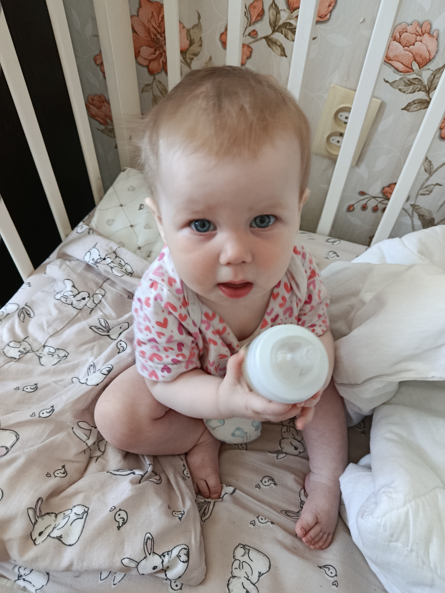 A baby with blue eyes and fair skin sits in a crib holding a white baby bottle. The baby wears a light-colored outfit with pink and red hearts. The crib has a patterned sheet with drawings of rabbits and birds. The crib's railings are white, and the wallpaper behind the crib features floral patterns. A European-style electrical outlet is visible on the wall behind the crib. The crib appears to be indoors. There is no indication of the city Фоки.
