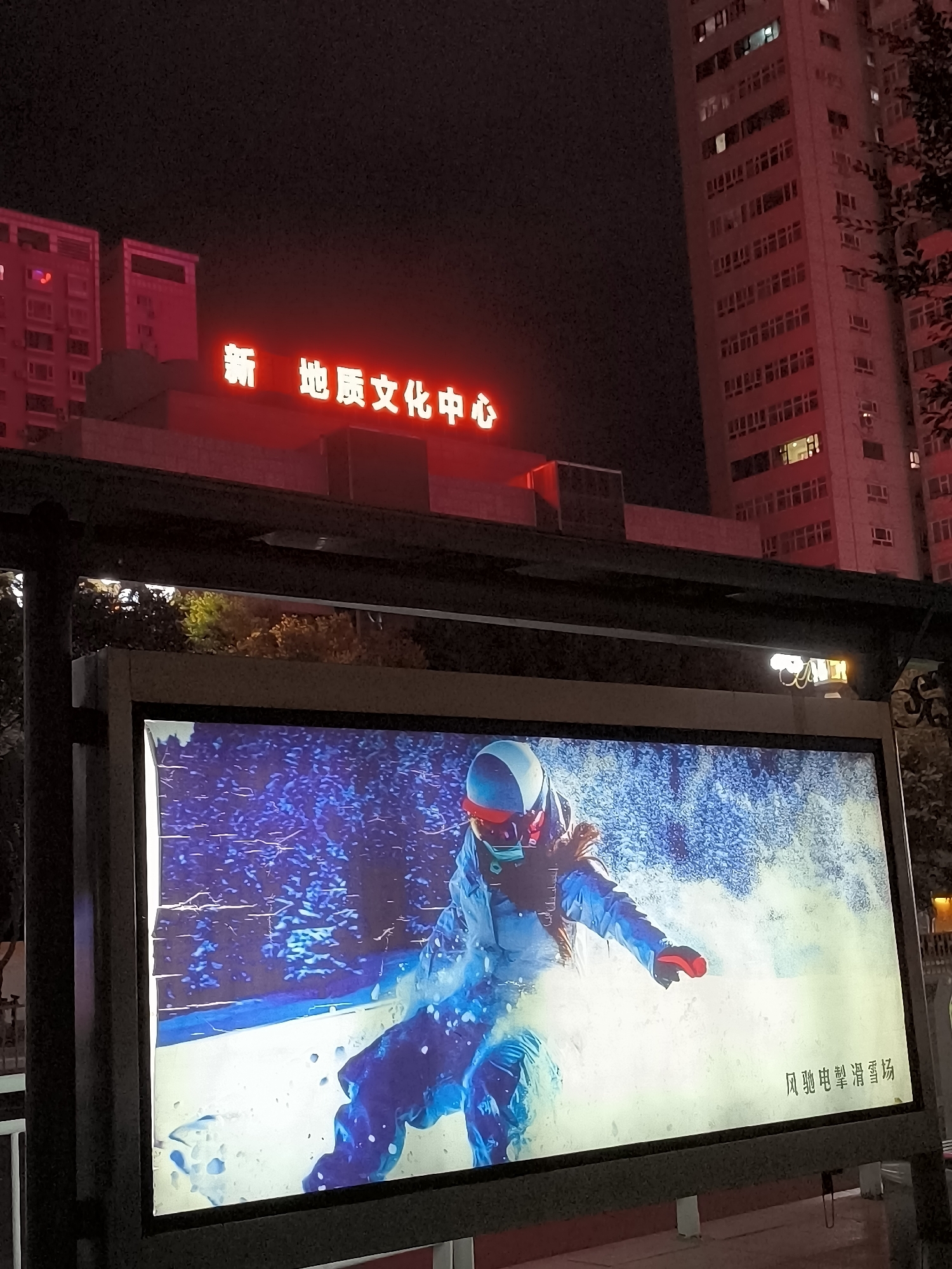 The image shows a nighttime scene in Ürümqi, China, featuring a red neon sign that reads "新 地质文化中心" (New Geological Cultural Center) above a bus stop shelter. The shelter has a lit advertising display showing a snowboarder in action against a blue, snowy background. To the right of the bus stop, a tall, multi-story building with many illuminated windows is visible. Trees are also present in the scene, some with visible foliage. The overall atmosphere is urban and nocturnal, with the neon sign and building lights providing illumination.