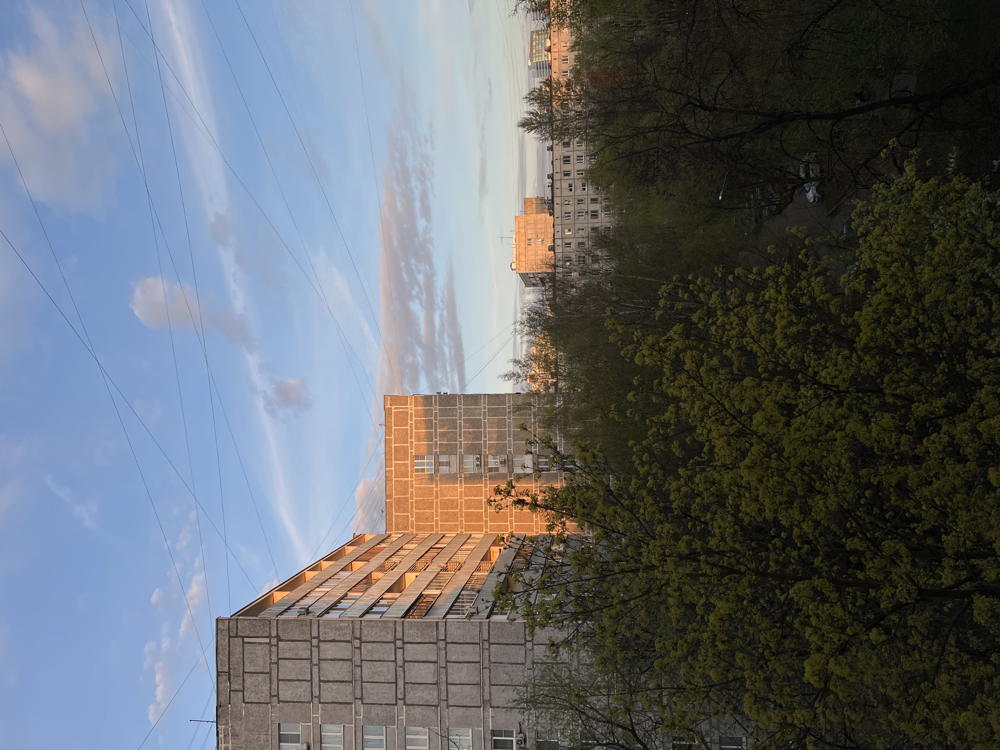 The image shows a residential area in Khimki, Russia, under a partly cloudy sky with electrical wires visible against the blue sky. In the foreground are Soviet-era apartment buildings with a precast concrete facade. Some of the apartments on one building have balconies and windows are reflecting the warm glow of the setting sun. Lush green trees are adjacent to the building. Distant apartment buildings are visible through the trees.