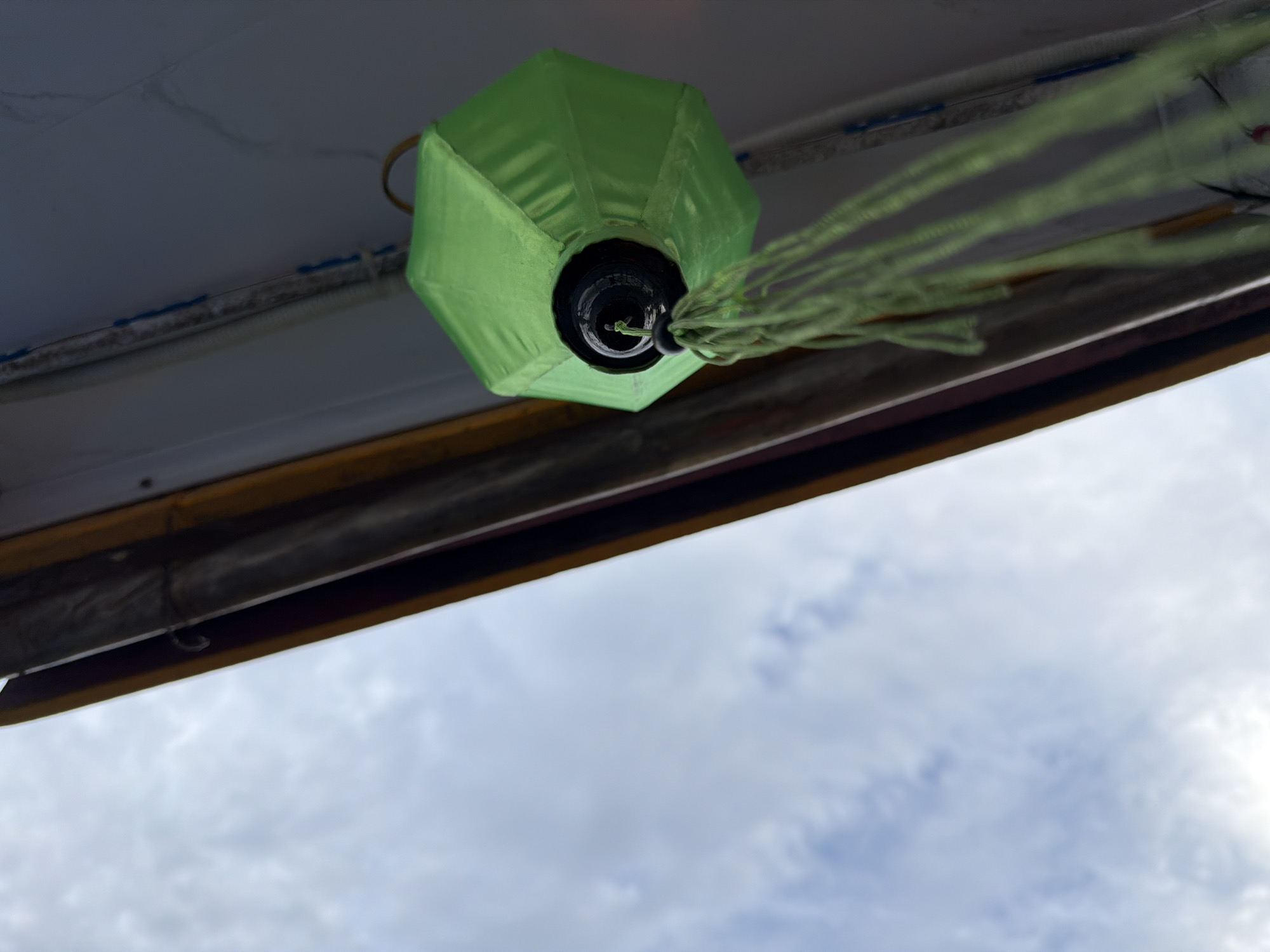 An upward-facing, low-angle shot captures a vibrant green lantern hanging from an overhead structure against a cloudy sky. The main subject is a hexagonal or octagonal fabric lantern, bright green in color, suspended from a dark, cylindrical fixture. Multiple light green tassels or strings extend from the bottom of the lantern, appearing to flow to the right, suggesting either motion or a gentle breeze.
The lantern is affixed to a white, slightly textured overhead surface, which could be the underside of a ceiling, a boat canopy, or an awning. Below this white surface, a dark horizontal beam or edge runs across the middle of the frame, featuring a distinct, narrow yellow stripe along its lower side. This structural element provides context, hinting at a built environment, possibly a vessel or a building with an overhang.
The lower half of the image consists of an overcast sky, filled with soft, diffused white and light grey clouds. This indicates a daytime setting, likely cloudy or before sunset, rather than bright sunshine. There are no people or specific activities depicted; the scene is static, focusing on the architectural detail and the decorative lantern. The setting suggests an outdoor or semi-outdoor environment, common in coastal or riverine locations like Đà Nẵng, Vietnam. No visible text is present.